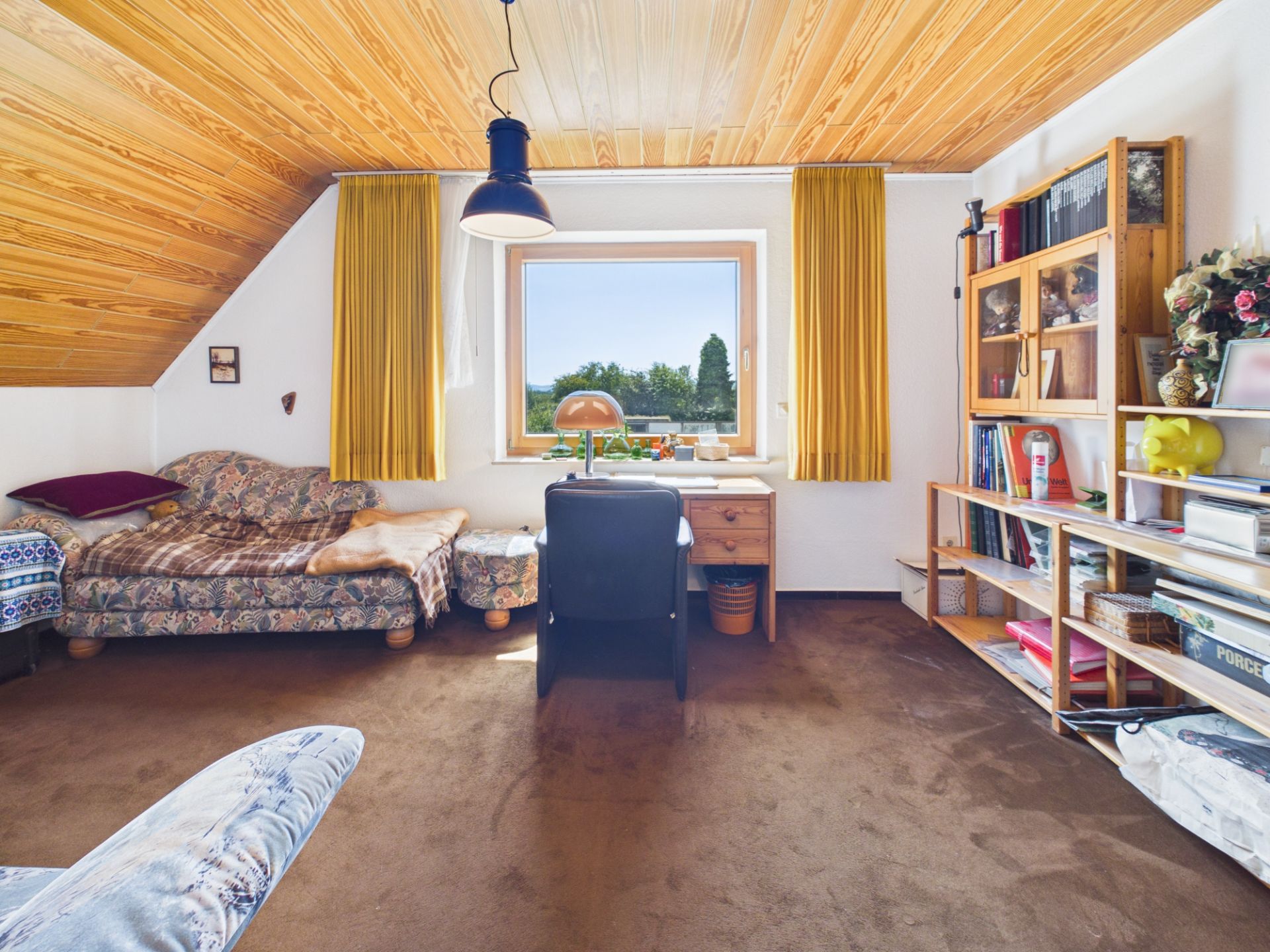 Helles Zimmer mit Schreibtisch, Regal, Sofa, gelben Vorhängen und Holzdecke.
