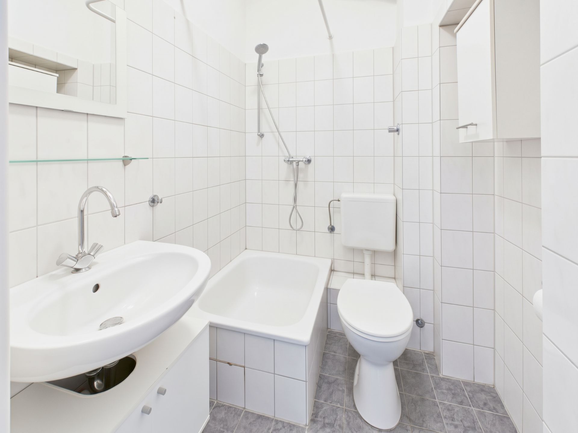 Kleines, weiß gefliestes Bad mit Waschbecken, Badewanne, Toilette und Dusche.