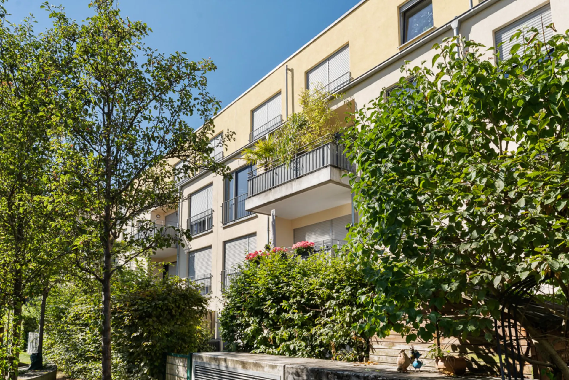 Mehrfamilienhaus mit Balkonen und viel grüner Bepflanzung im Vordergrund, sonniger Tag.