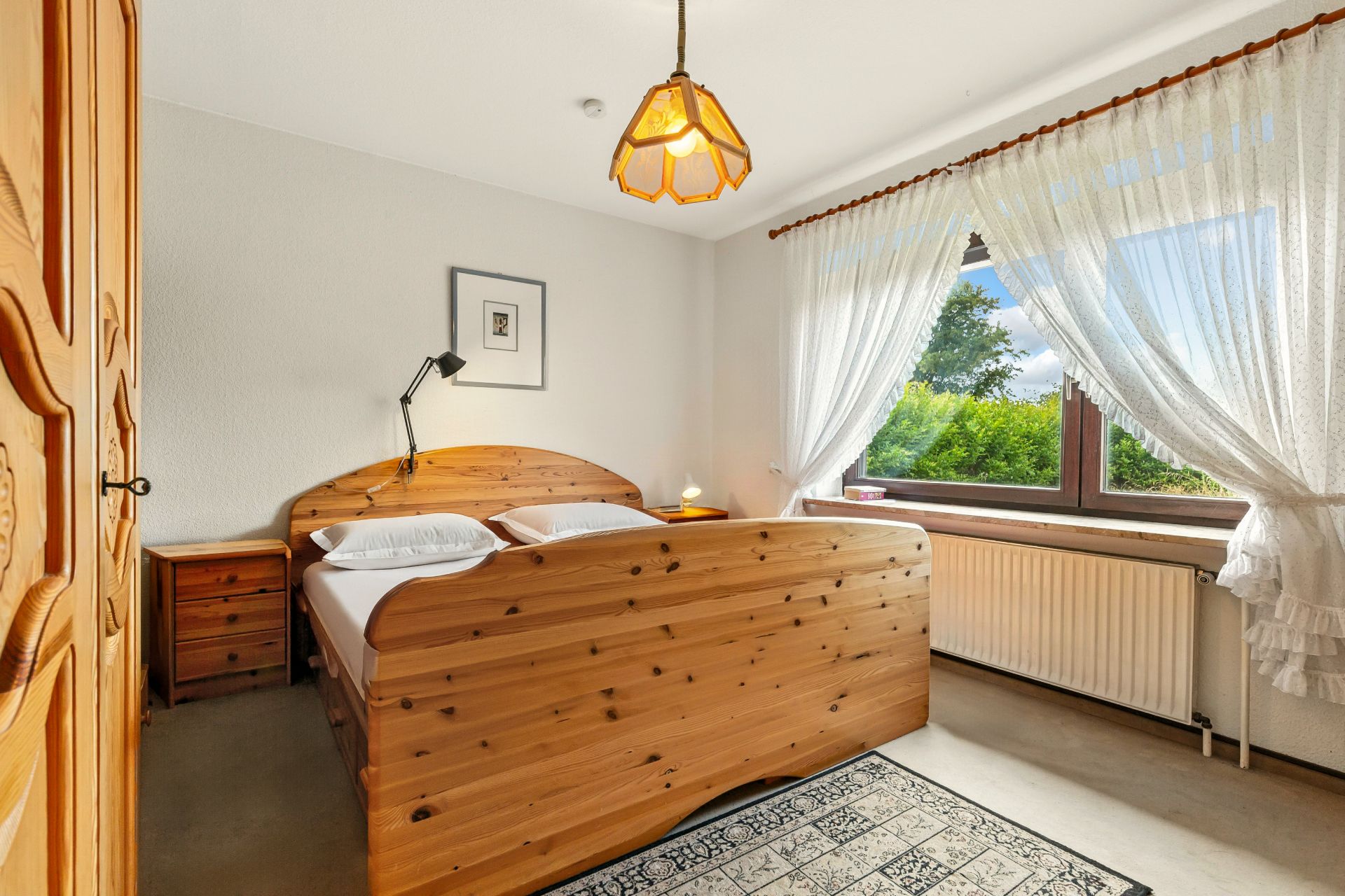 Schlafzimmer mit Holzbett, Nachttischen, Teppich und großem Fenster mit weißen Vorhängen.