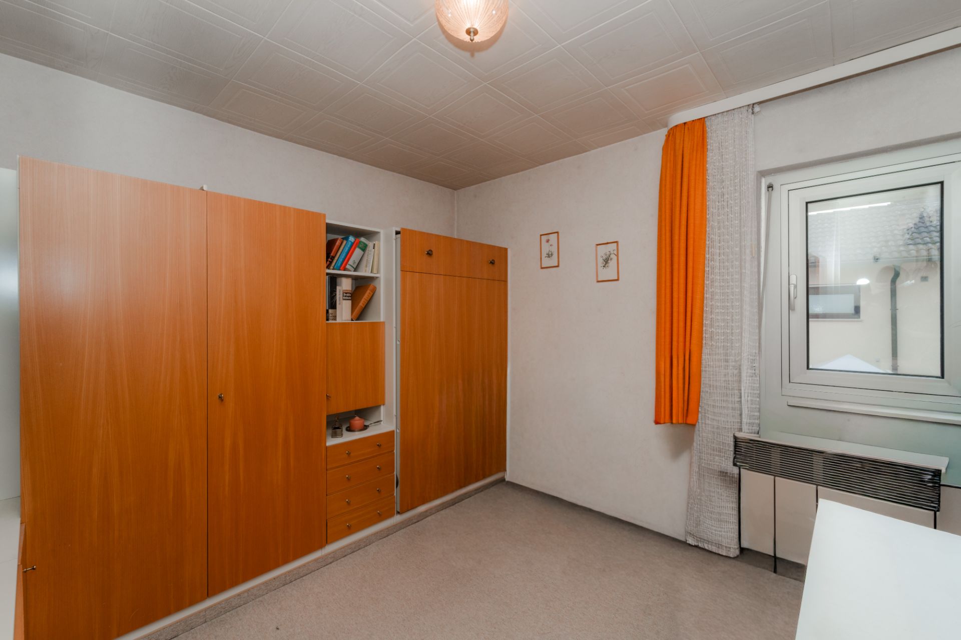 Schlichtes Zimmer mit Holzschrank, Regal, Fenster, oranger Gardine und Heizung.