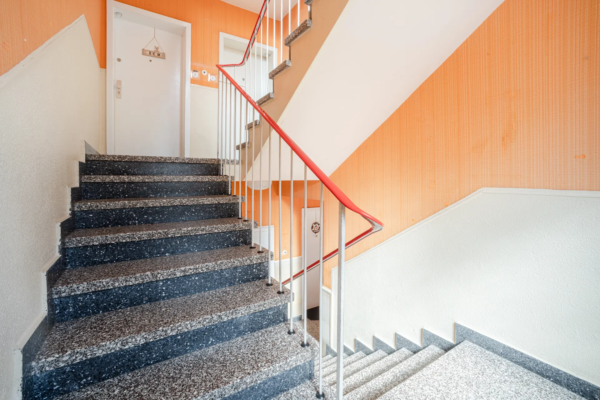 Treppenhaus mit orangefarbener Wand, grauen Stufen und weißer Tür mit Willkommensschild.