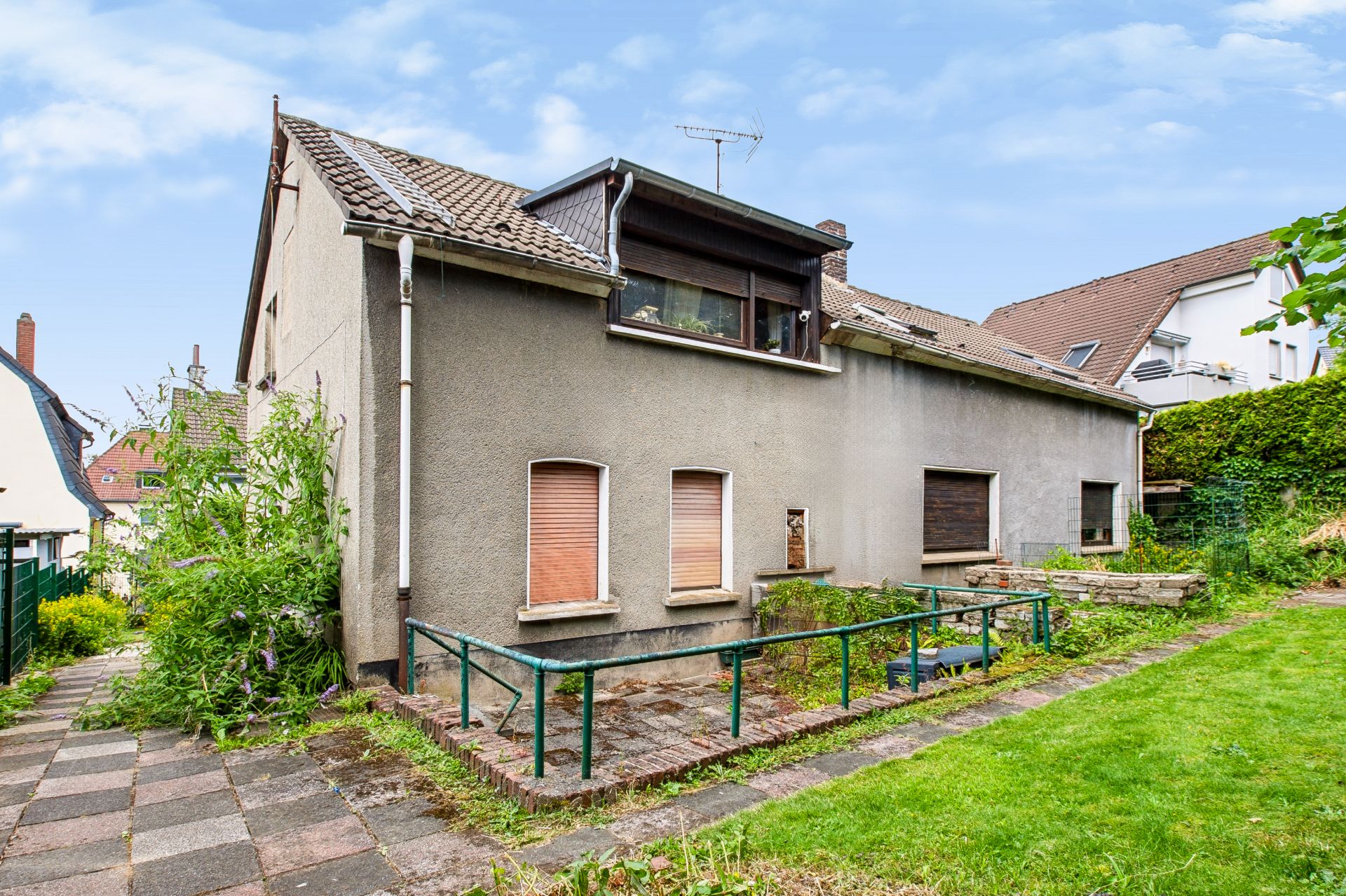 Graues Wohnhaus mit Rampe, geschlossenen Fensterläden und Garten im Vordergrund.