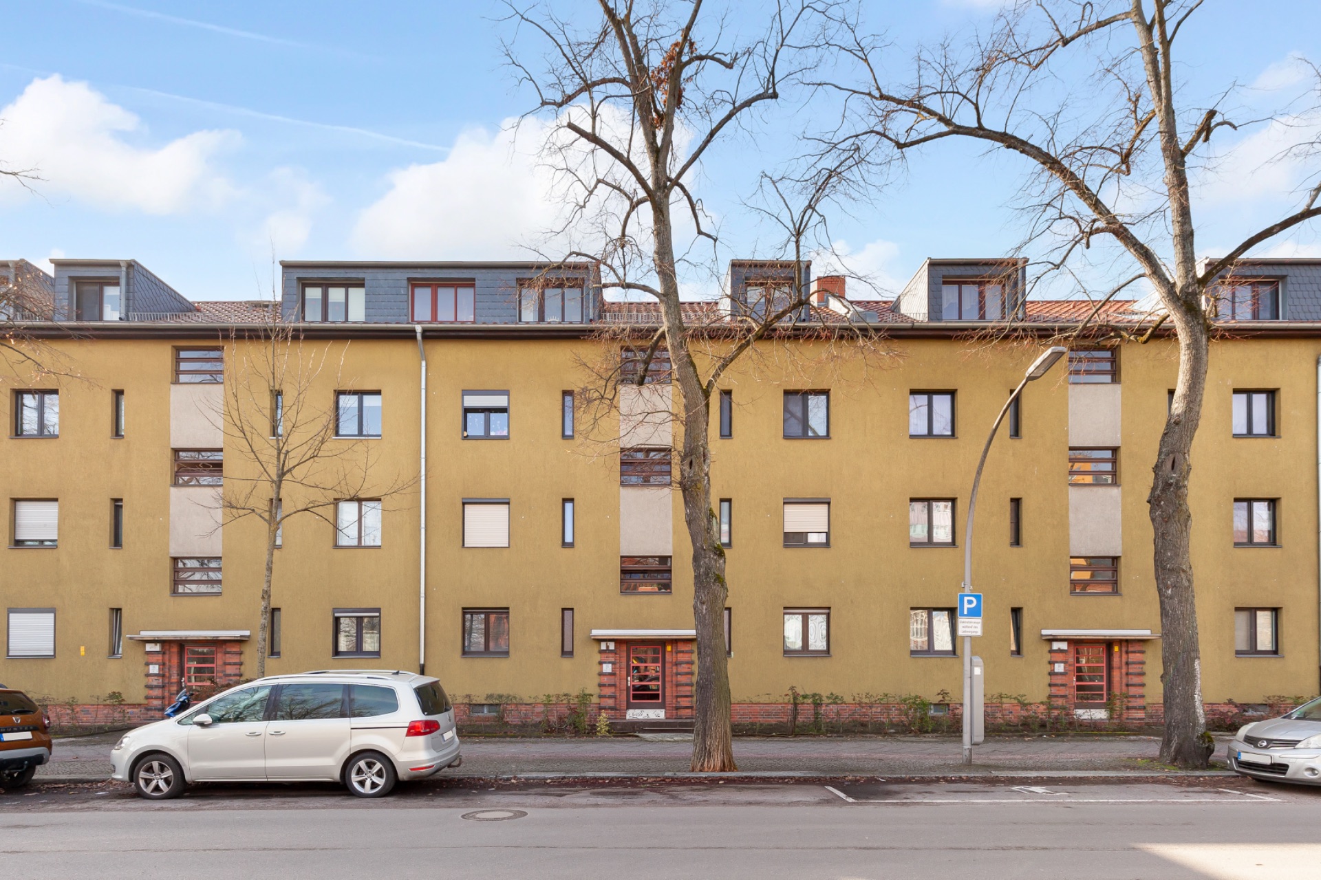 Mehrfamilienhaus mit gelber Fassade, mehreren Eingängen, Bäumen und geparkten Autos davor.