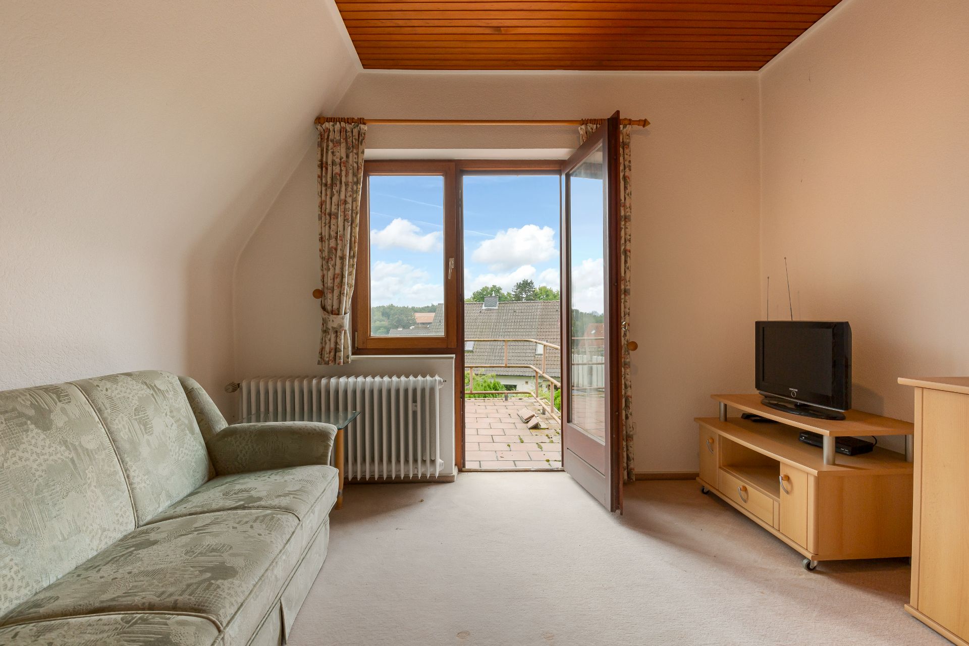 Wohnzimmer mit grünem Sofa, Fernseher auf Holzregal und Tür zu einem Balkon mit Blick nach draußen.