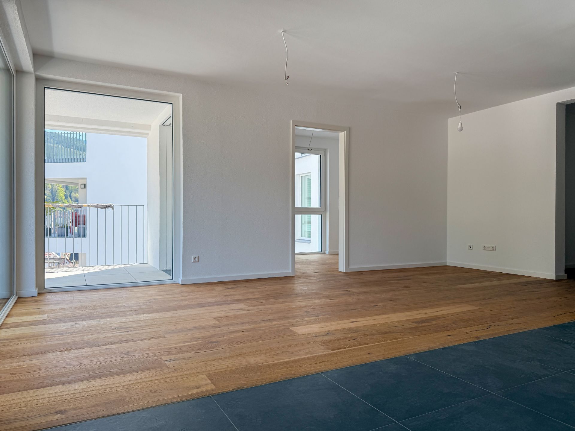 Helles, leeres Wohnzimmer mit Holzboden, Fliesenbereich und Zugang zu einem Balkon.