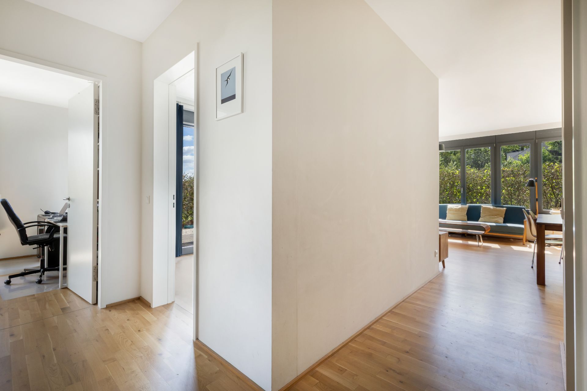 Heller Flur mit Holzboden, Zugang zu Arbeitszimmer und Wohnzimmer mit Sofa und großen Fenstern.