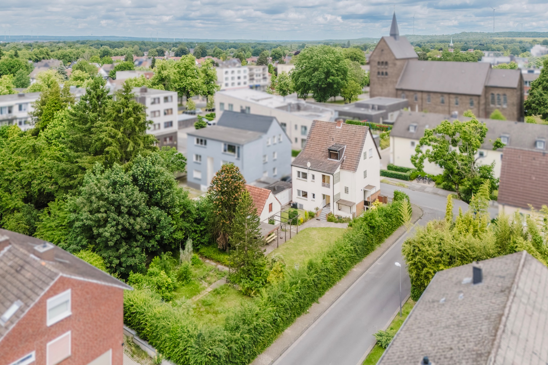 Luftaufnahme eines Wohngebiets mit Einfamilienhäusern, Bäumen und einer Kirche im Hintergrund.