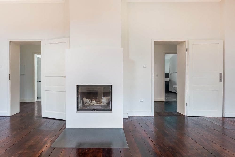 Helles Zimmer mit Kamin, dunklem Holzboden und zwei offenen weißen Türen.