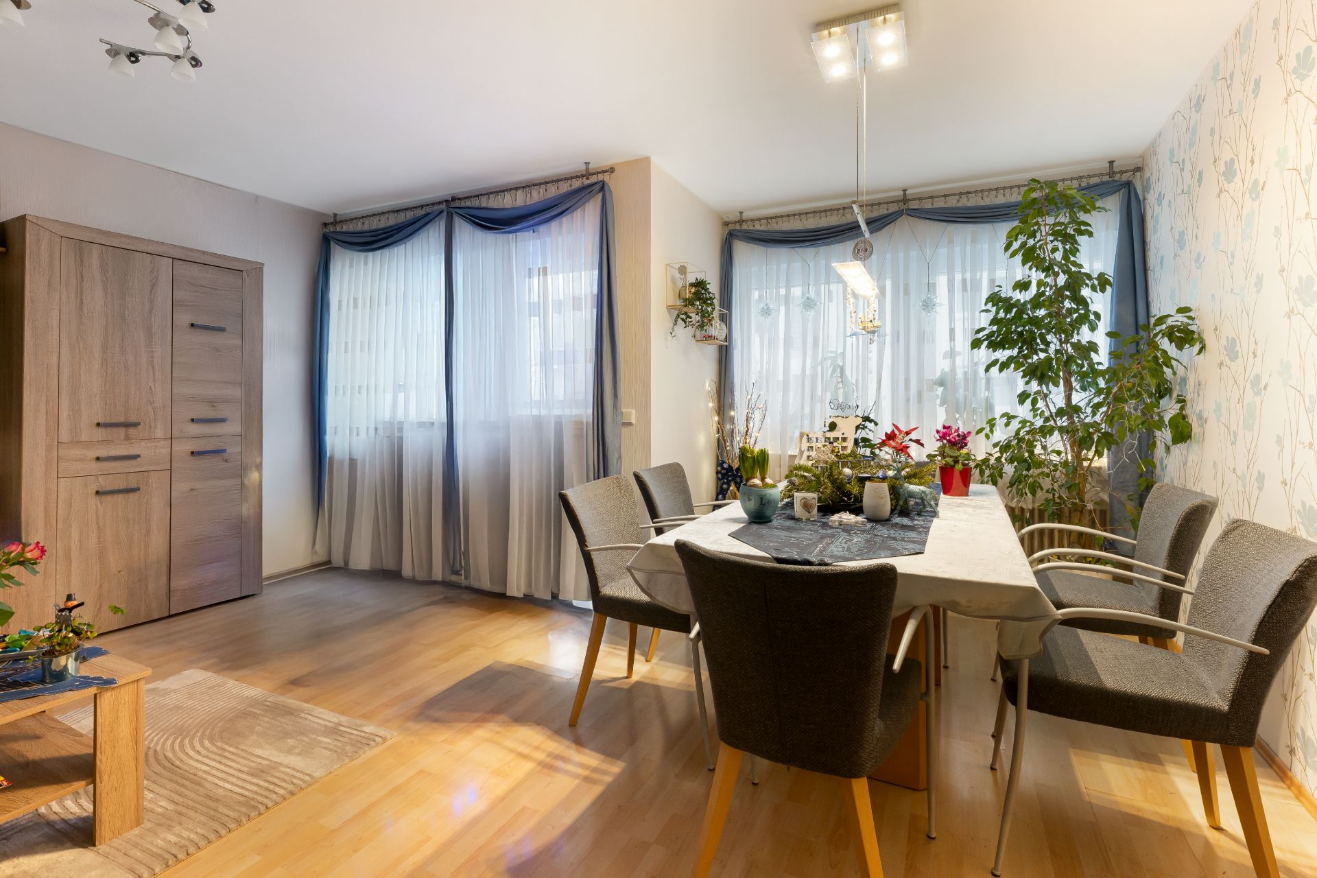 Essbereich mit Holztisch, sechs Stühlen, Pflanzen und großen Fenstern mit Vorhängen.