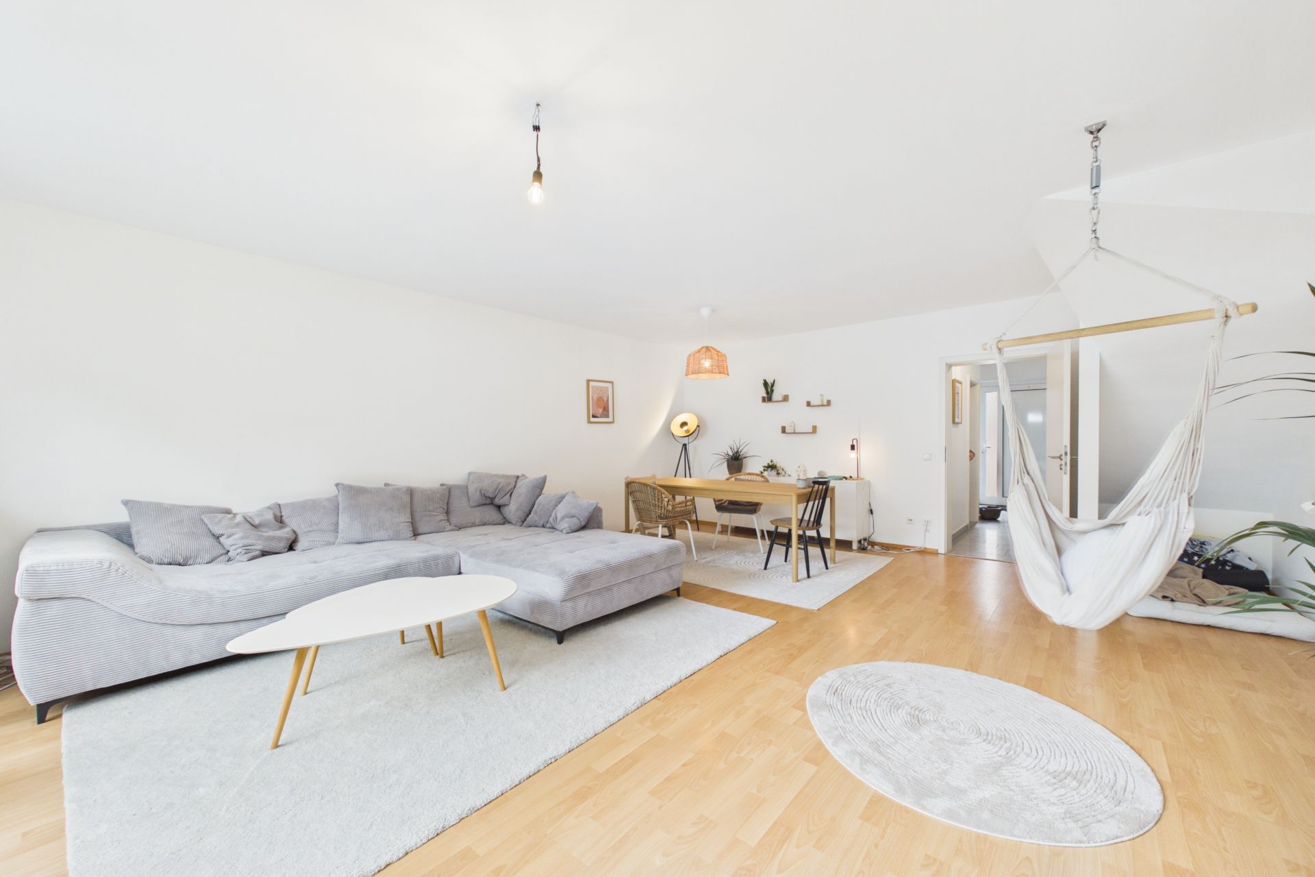 Helles Wohnzimmer mit grauem Sofa, Hängesessel, Esstisch und hellem Holzboden.