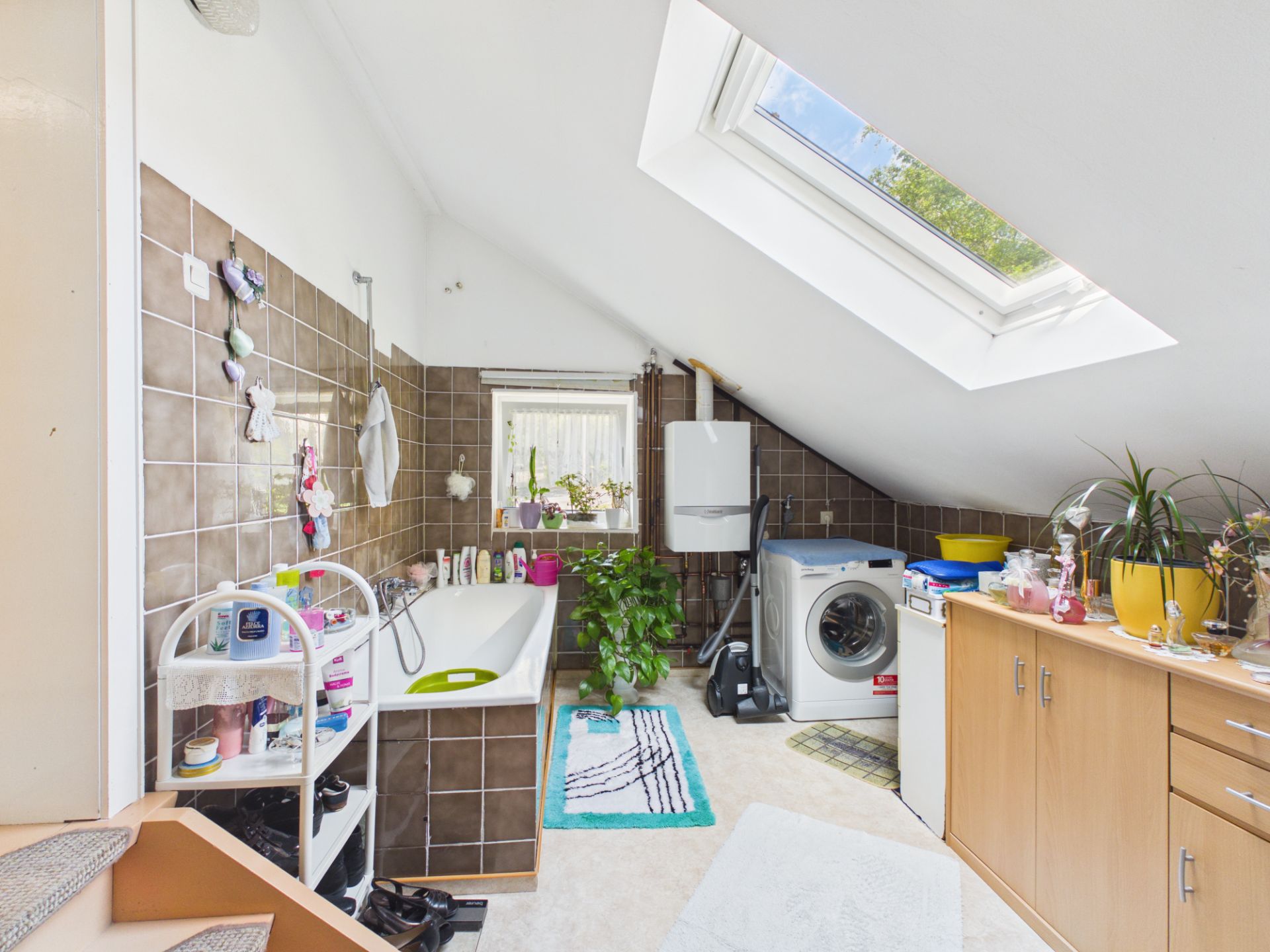 Helles Badezimmer mit Badewanne, Waschmaschine, Dachfenster und Pflanzen.