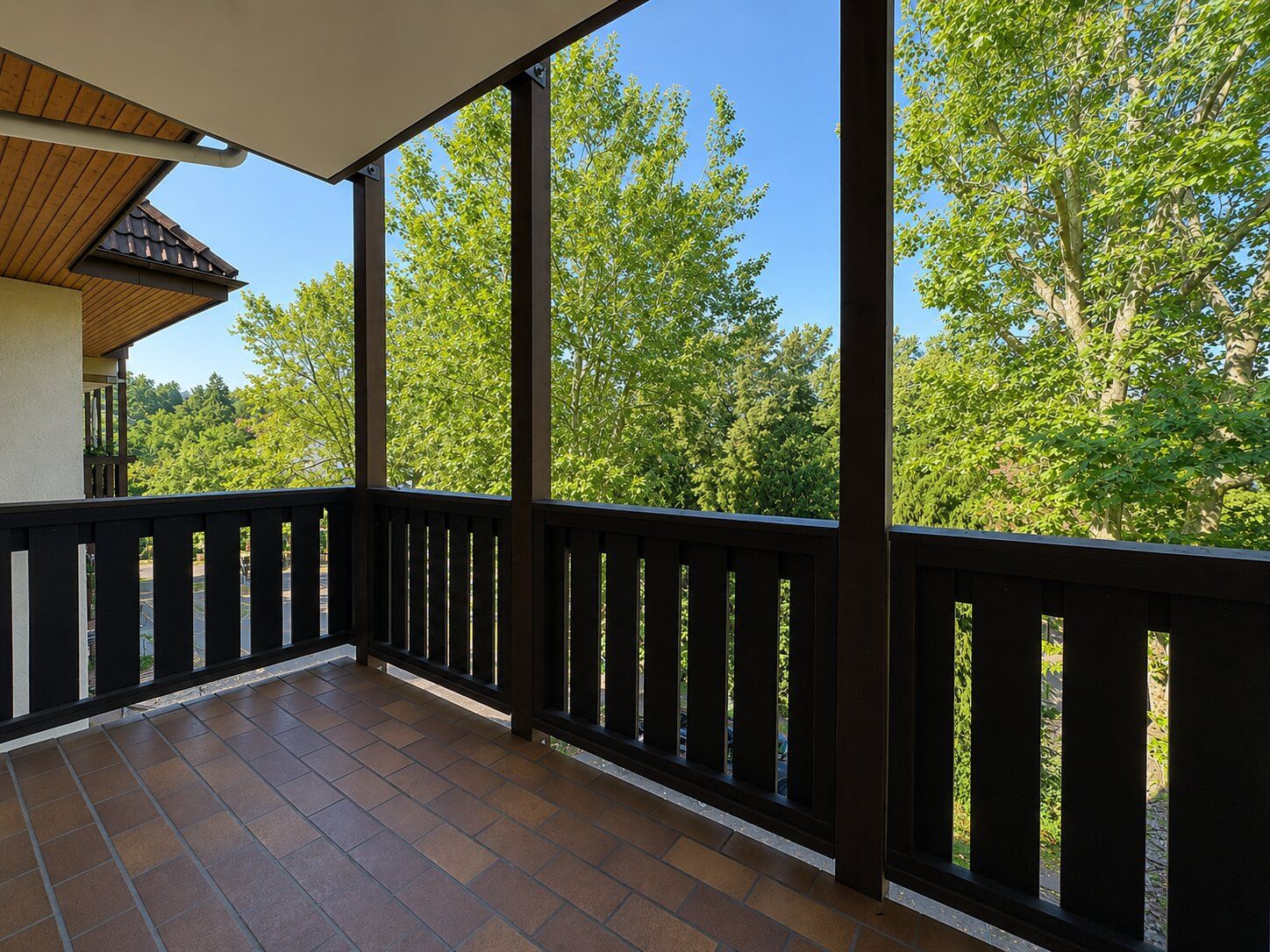 Überdachter Balkon mit braunem Geländer und Blick auf grüne Bäume.