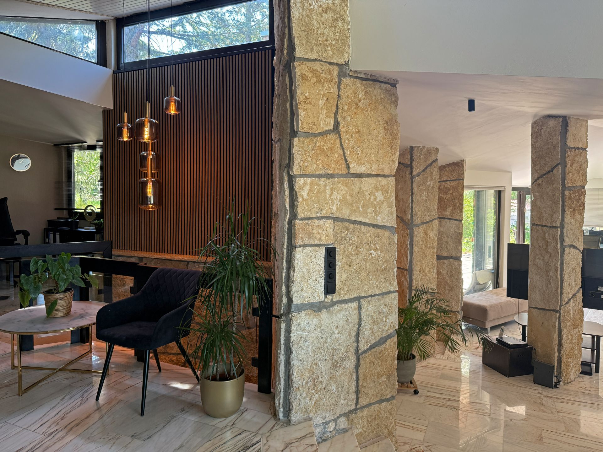 Wohnzimmer mit Naturstein-Säulen, Pflanzen, Sessel und Hängelampen, helle Fenster im Hintergrund.