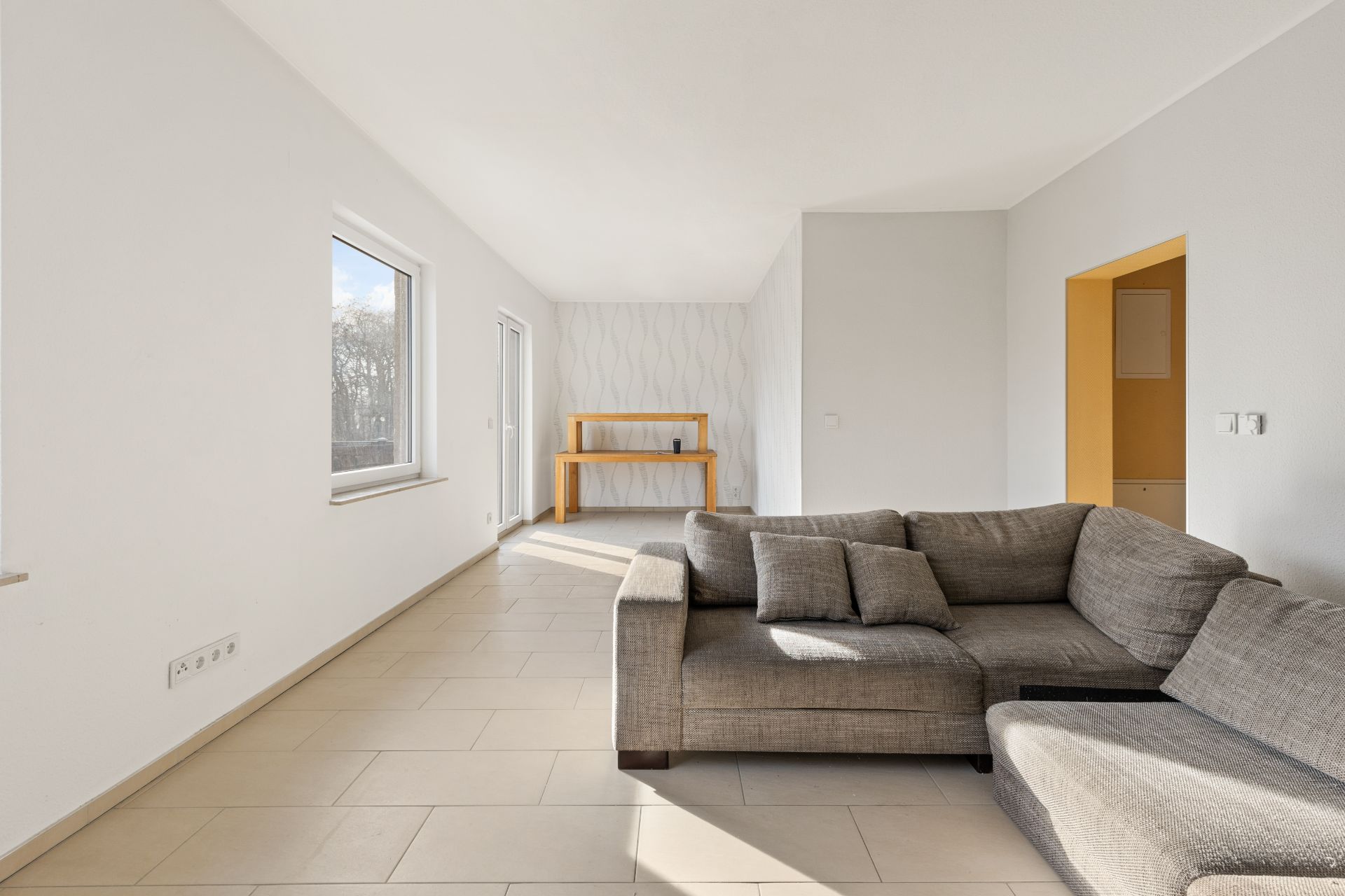 Helles Wohnzimmer mit grauem Sofa, großen Fenstern und einem Holztisch an der Wand.