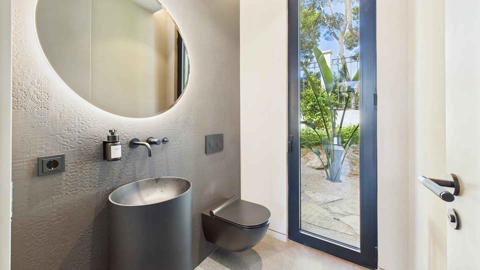 Modernes Badezimmer mit rundem Spiegel, schwarzem Waschbecken, Toilette und großem Fenster mit Gartenblick.