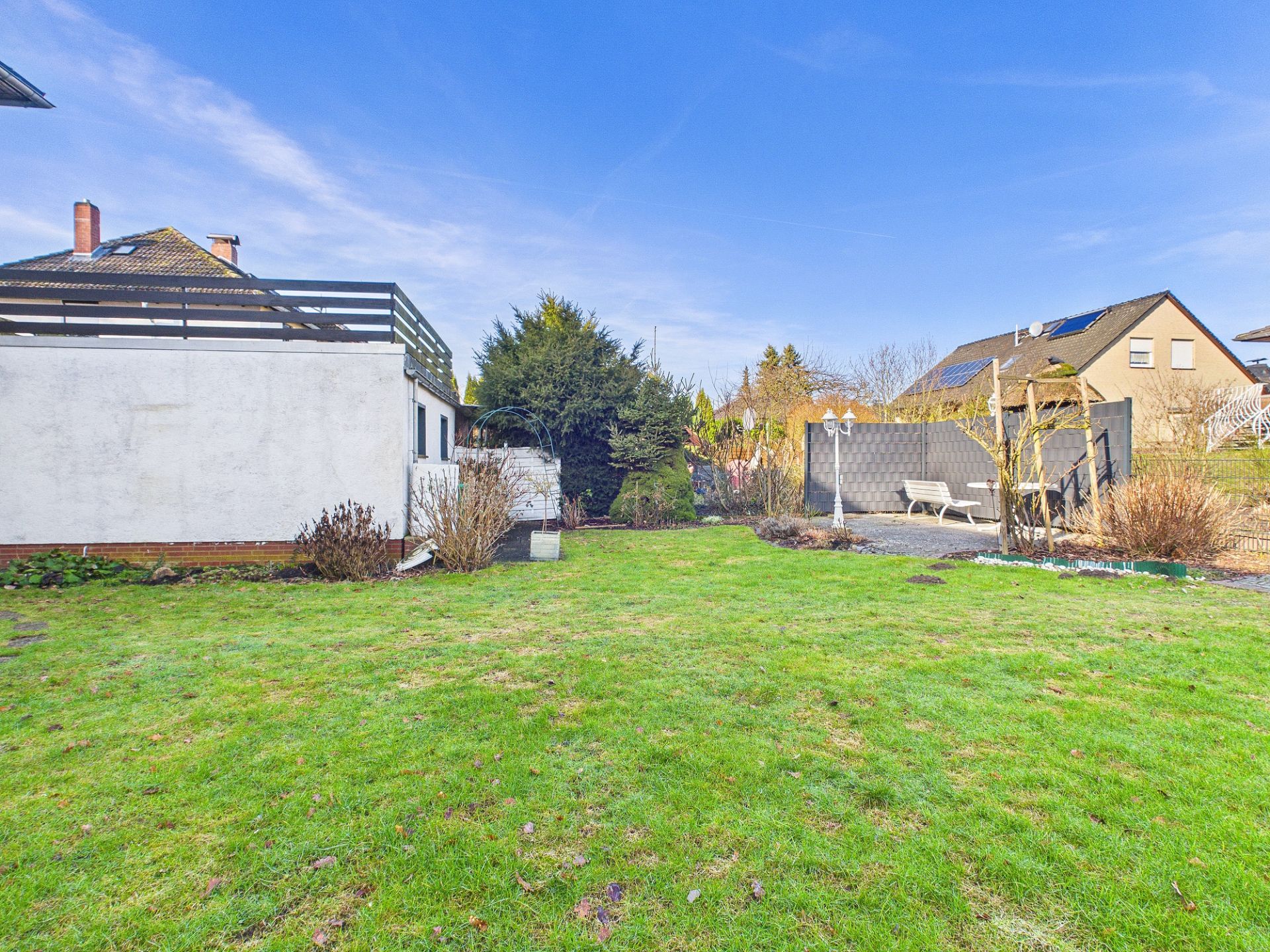 Großer Garten mit Rasen, Sträuchern, Bank und angrenzenden Wohnhäusern bei blauem Himmel.