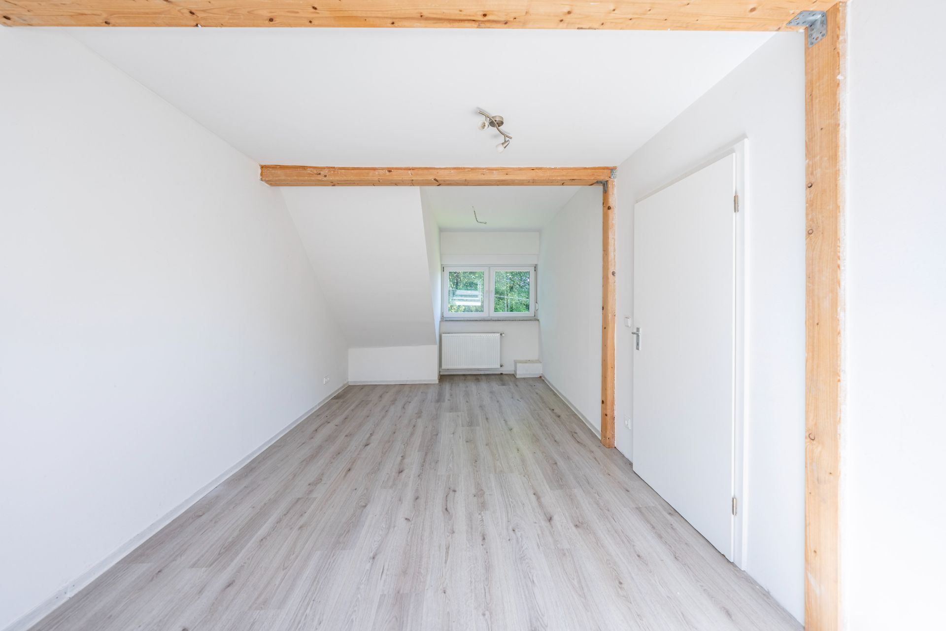 Heller, leerer Raum mit Holzrahmen, weißem Boden und Fenster am Ende.