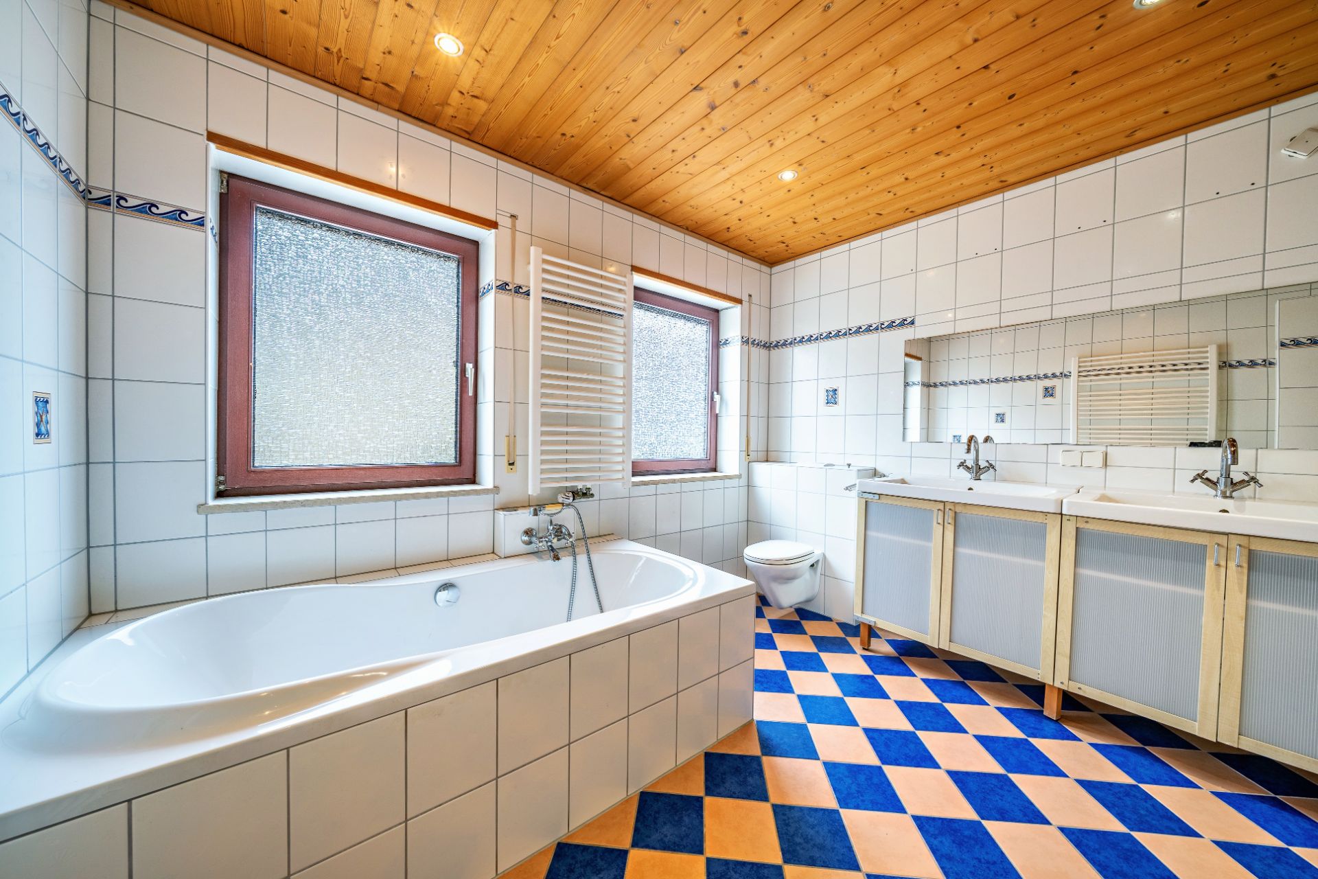 Helles Badezimmer mit Badewanne, Doppelwaschbecken, WC und Fenster, blau-orange karierten Fliesen.