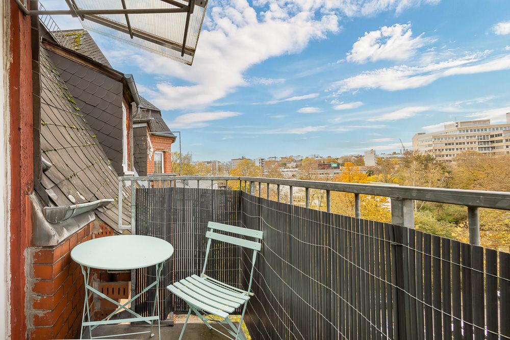 Kleiner Balkon mit Tisch und Stuhl, Blick auf Stadt und Bäume unter blauem Himmel.