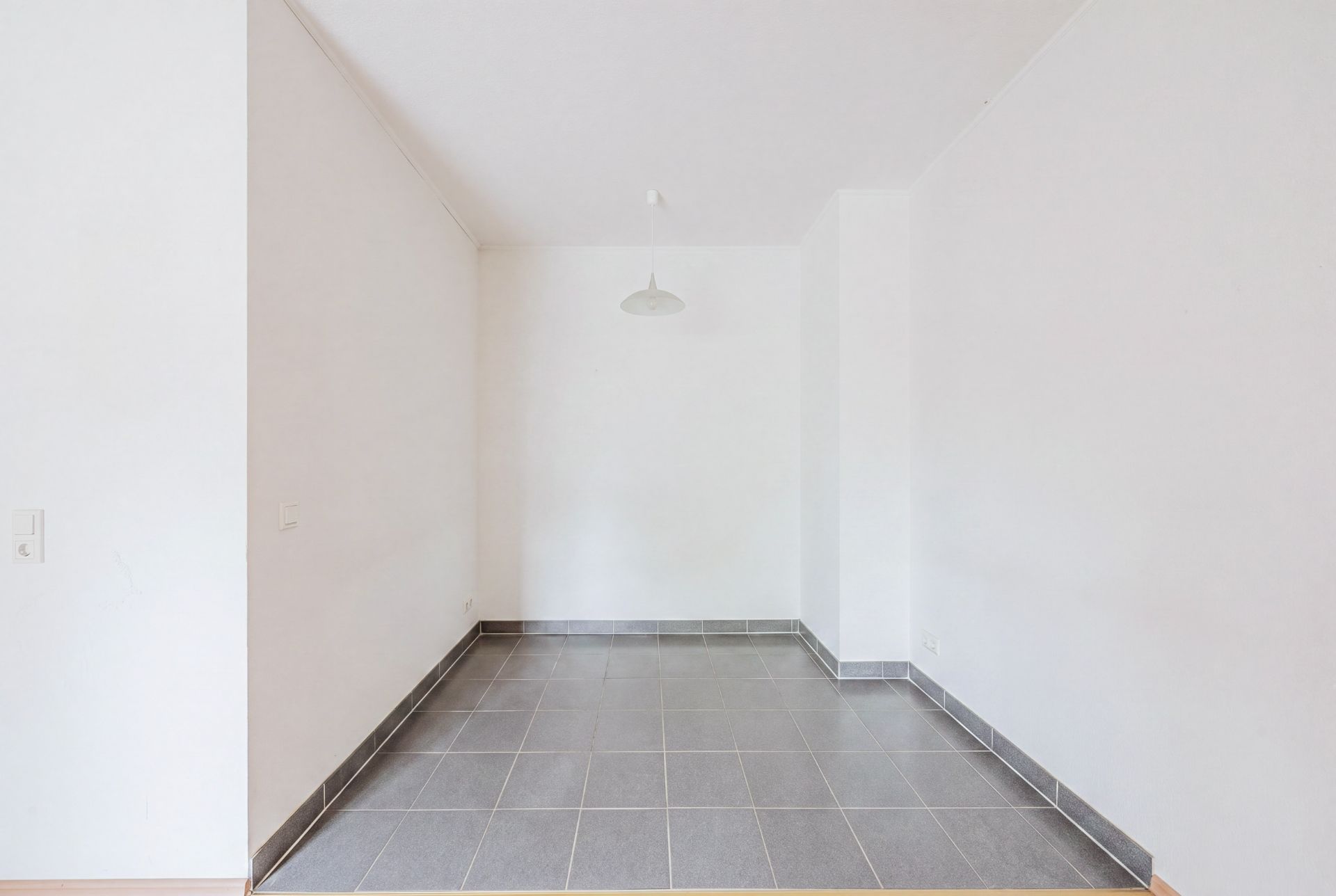 Leerer Raum mit grauen Fliesen und weiß gestrichenen Wänden, Deckenlampe in der Mitte.