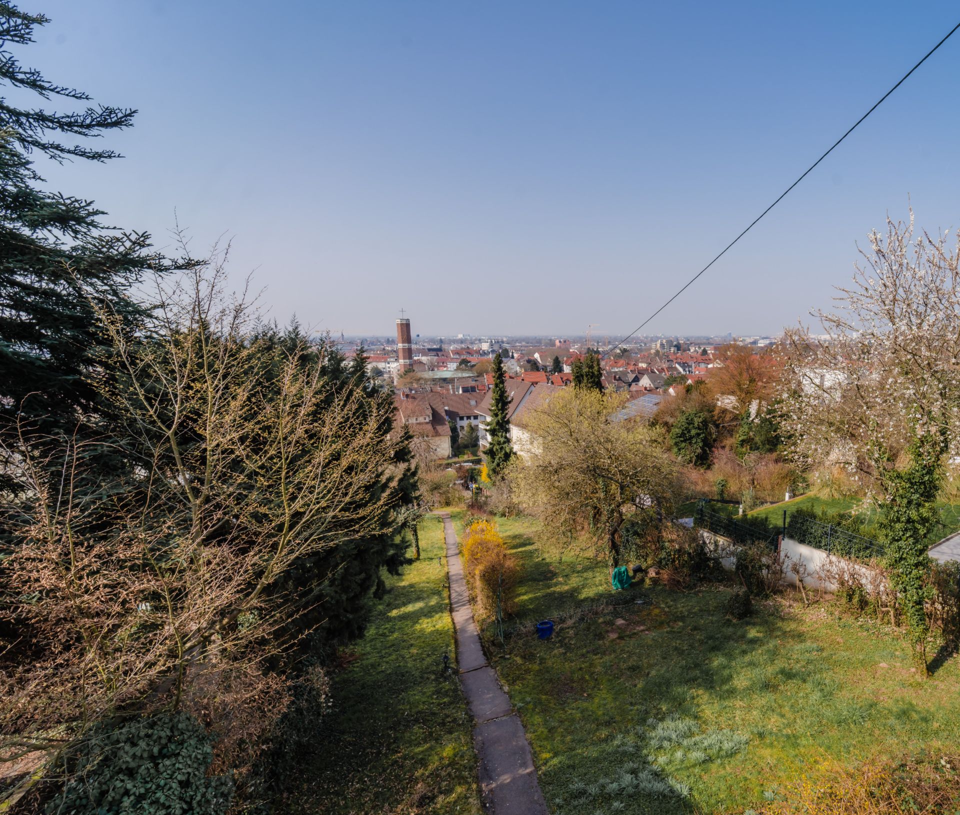 Blick über einen Garten mit Weg und Stadt im Hintergrund, Bäume an den Seiten.