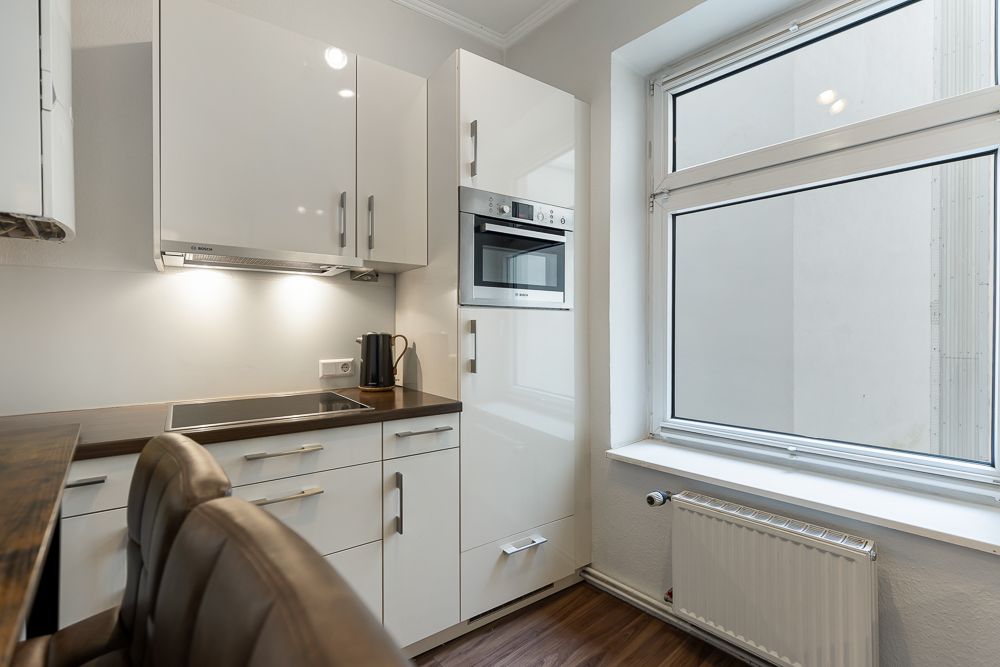 Moderne weiße Einbauküche mit Kochfeld, Backofen und großem Fenster.