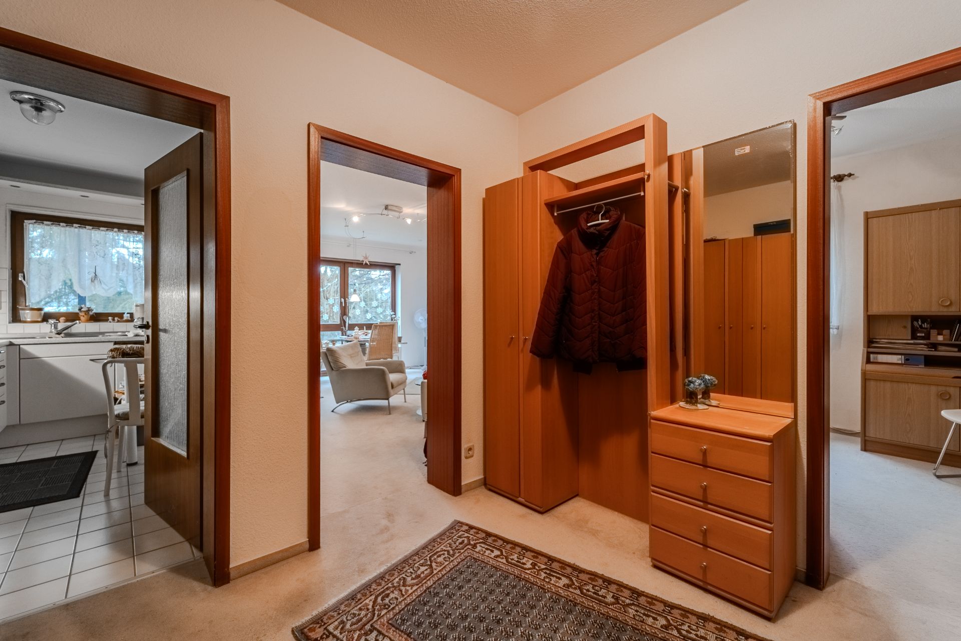 Flur mit Teppich, Garderobe, Spiegel und offenen Türen zu Küche, Wohnzimmer und Arbeitszimmer.