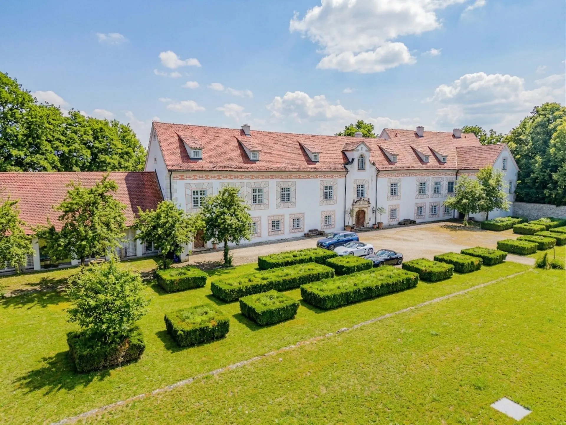 Großes weißes Wohngebäude mit rotem Dach, symmetrischem Garten und mehreren parkenden Autos vor dem Haus.