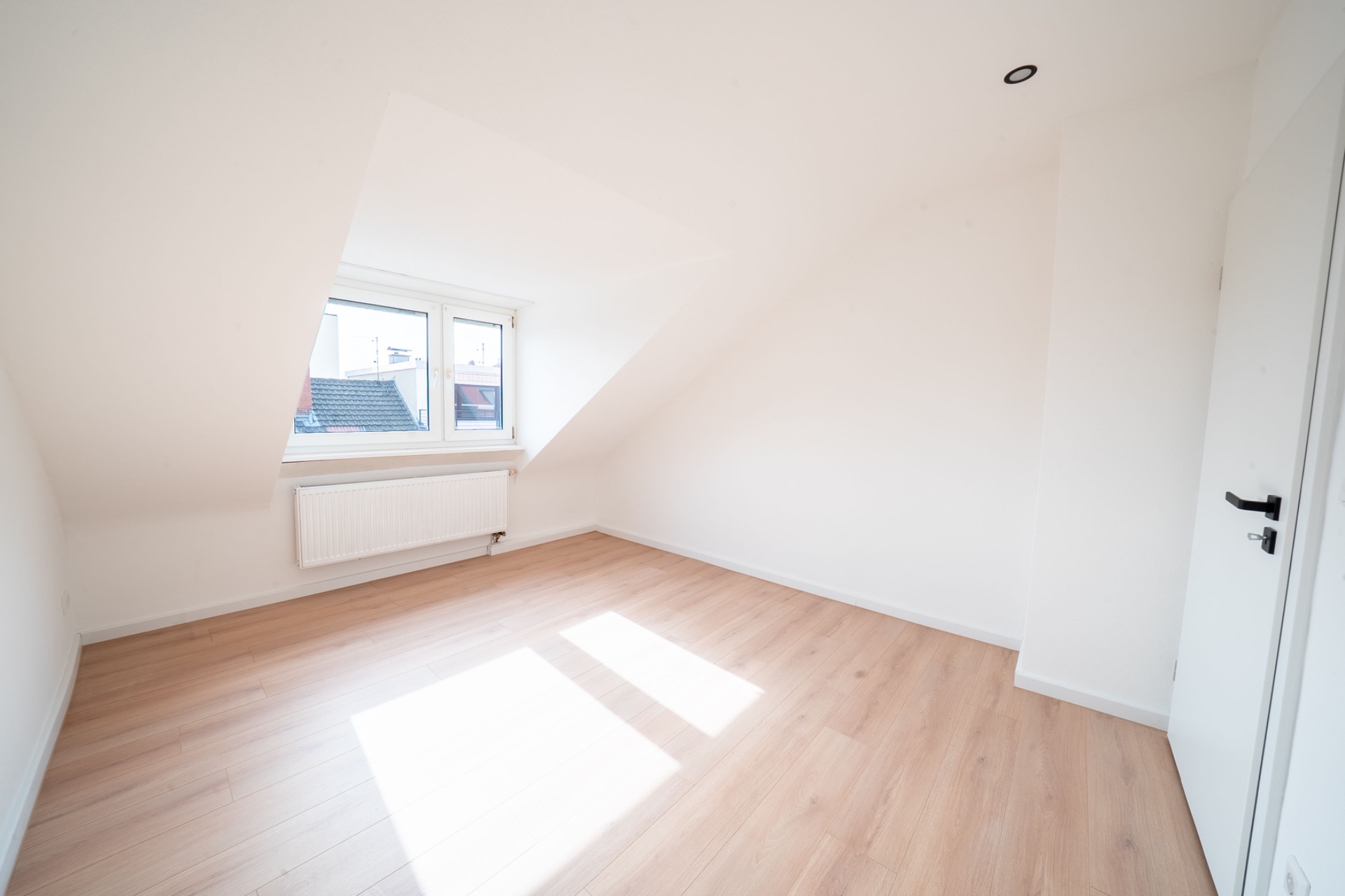 Helles, leeres Zimmer mit schräger Decke, großem Fenster und hellem Holzfußboden.