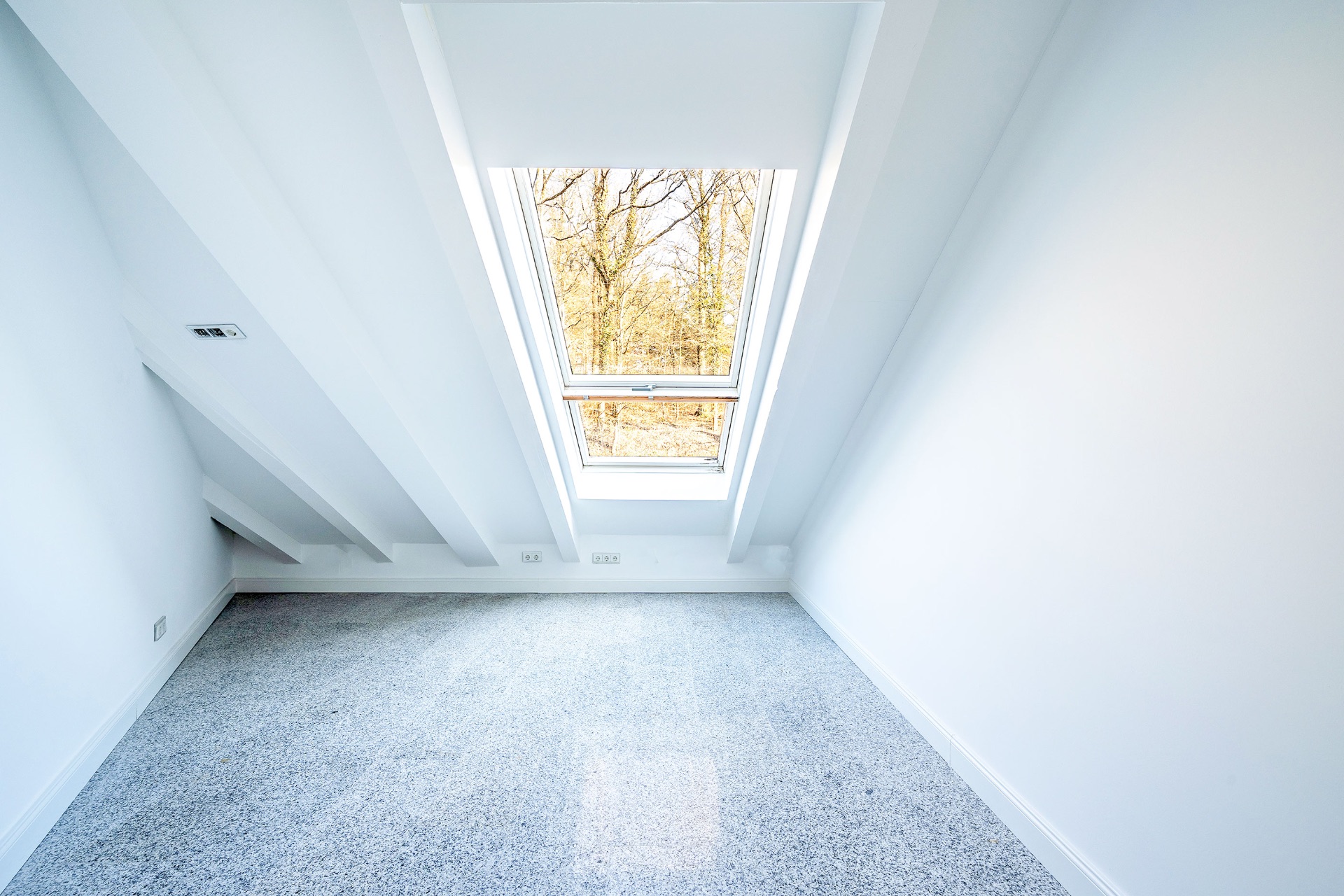 Heller Dachraum mit schrägen Wänden, großem Dachfenster und grauem Teppichboden.