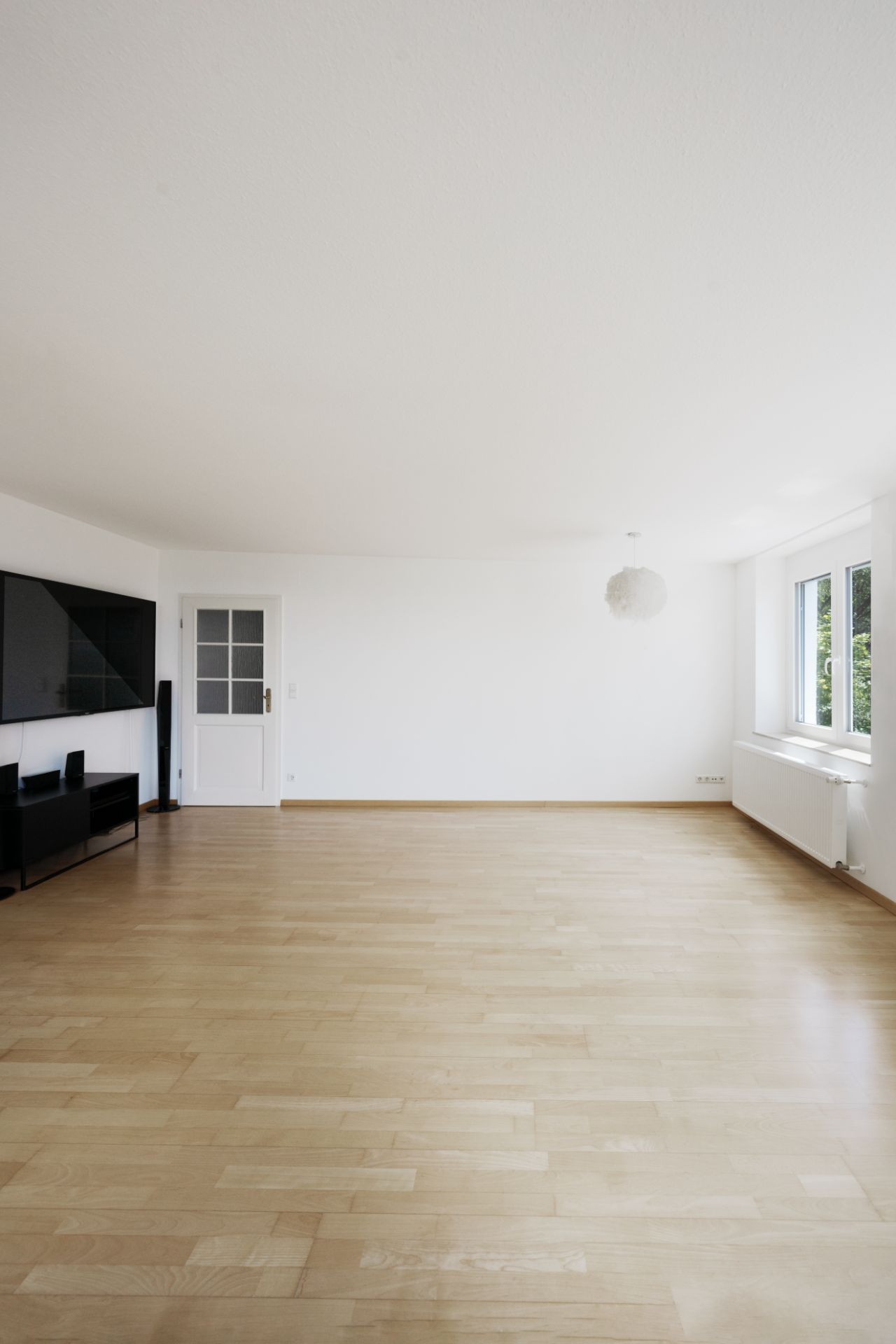 Helles, leeres Wohnzimmer mit Parkettboden, einem Fenster, weißer Tür und Fernseher an der Wand.