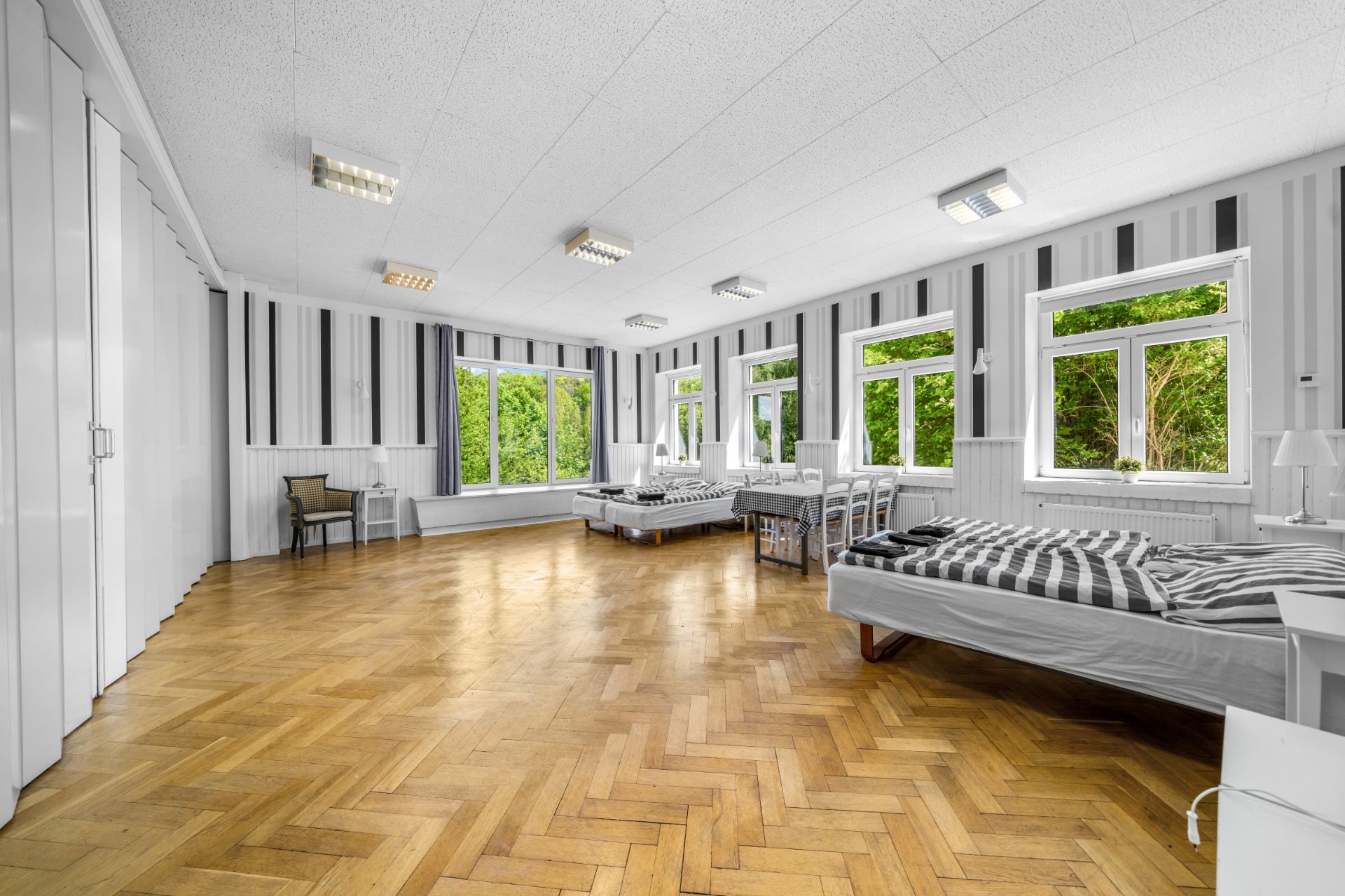 Großes Schlafzimmer mit mehreren Betten, Holzboden und großen Fenstern, die auf einen Garten blicken.