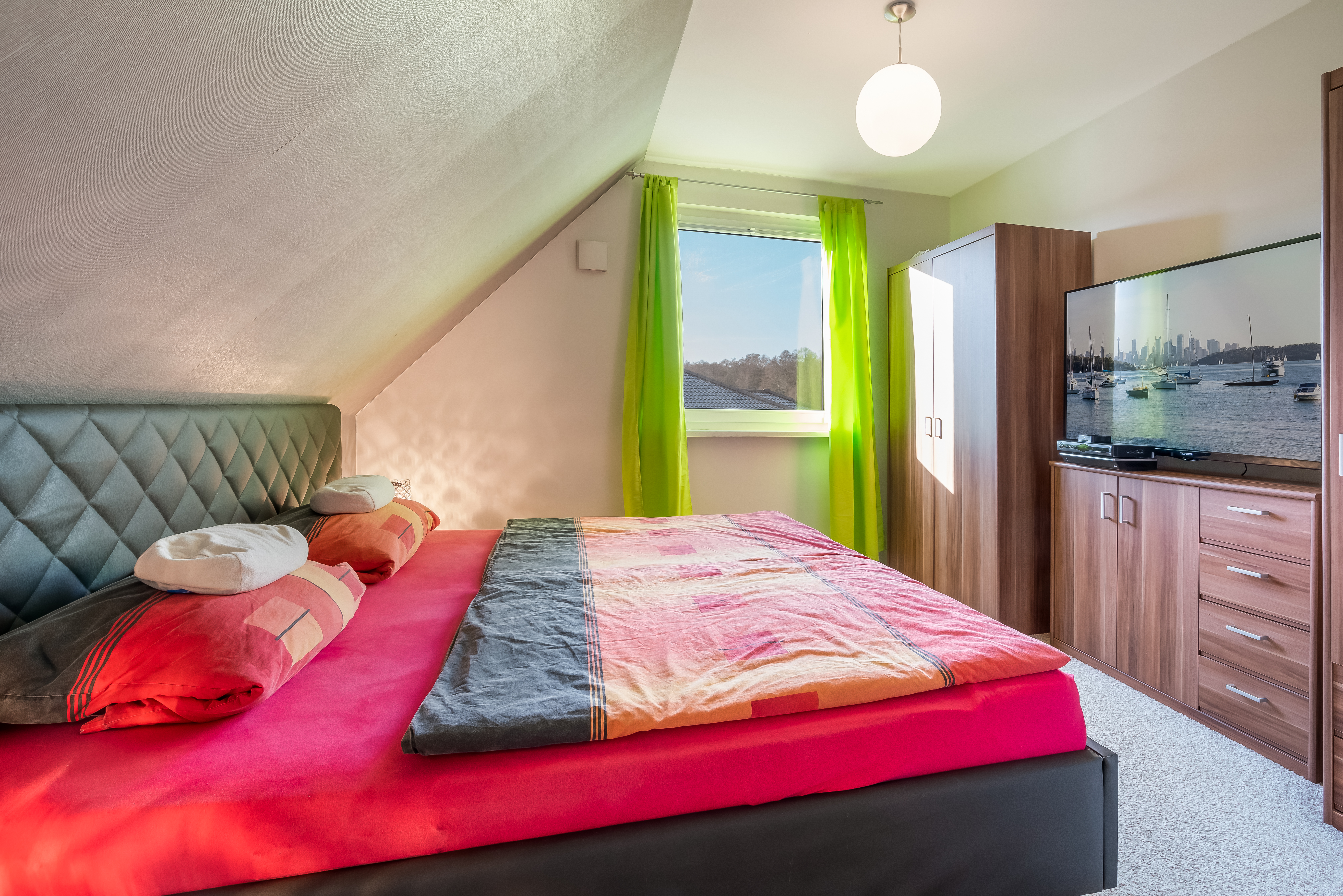 Helles Schlafzimmer mit Doppelbett, grünem Vorhang, Kleiderschrank und Fernseher auf einer Kommode.