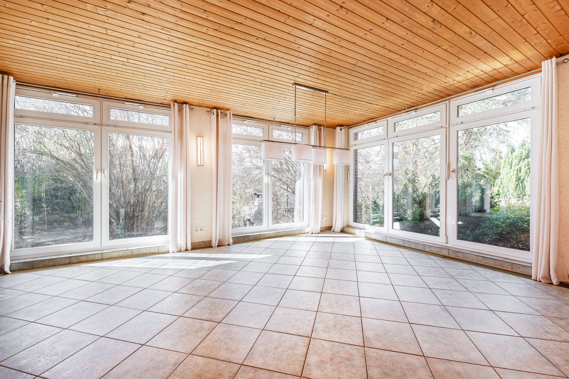 Heller Raum mit großen Fenstern, Fliesenboden und Holzdecke, Blick in den Garten.