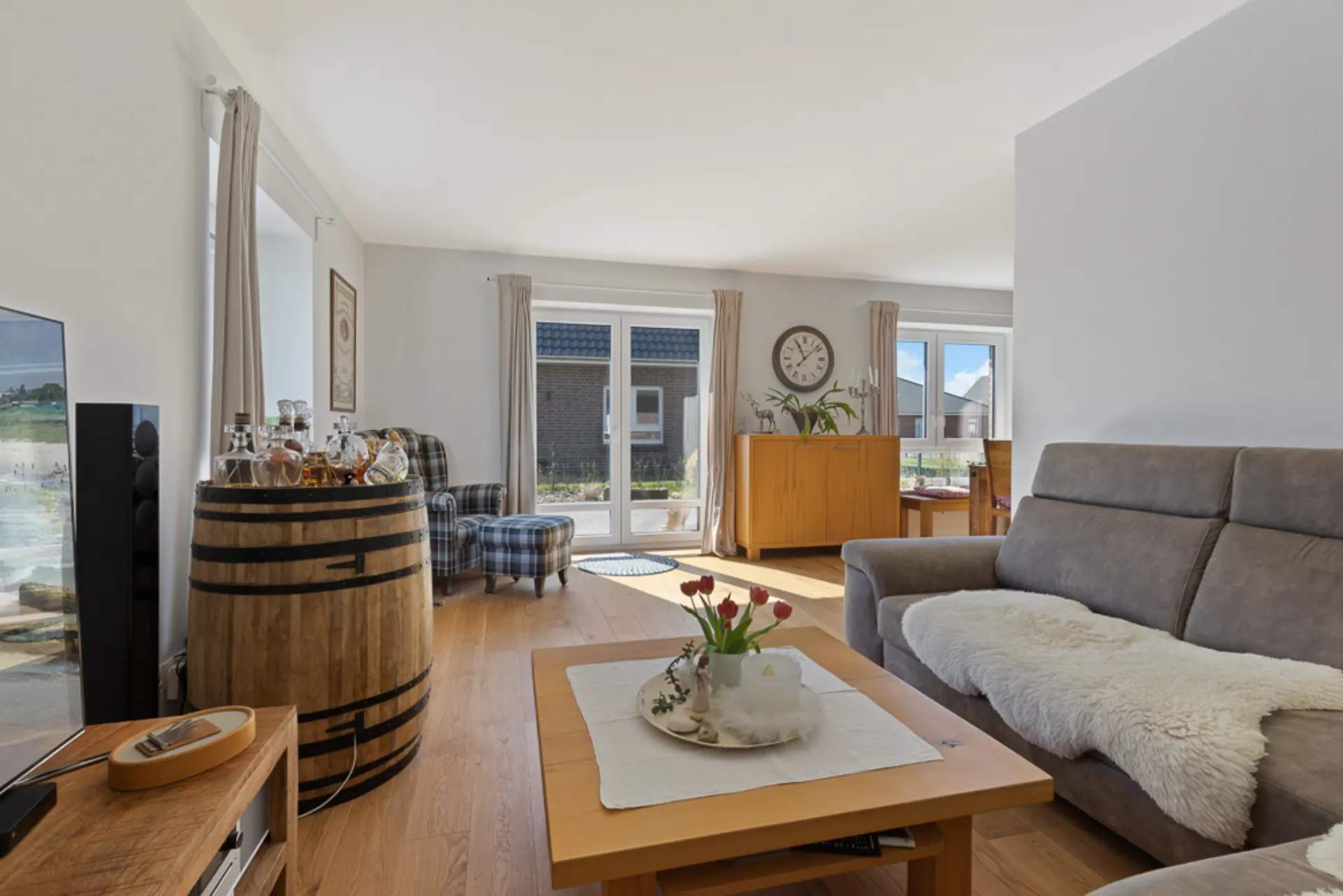 Helles Wohnzimmer mit grauem Sofa, Holztisch, Holzfass und großer Fensterfront.