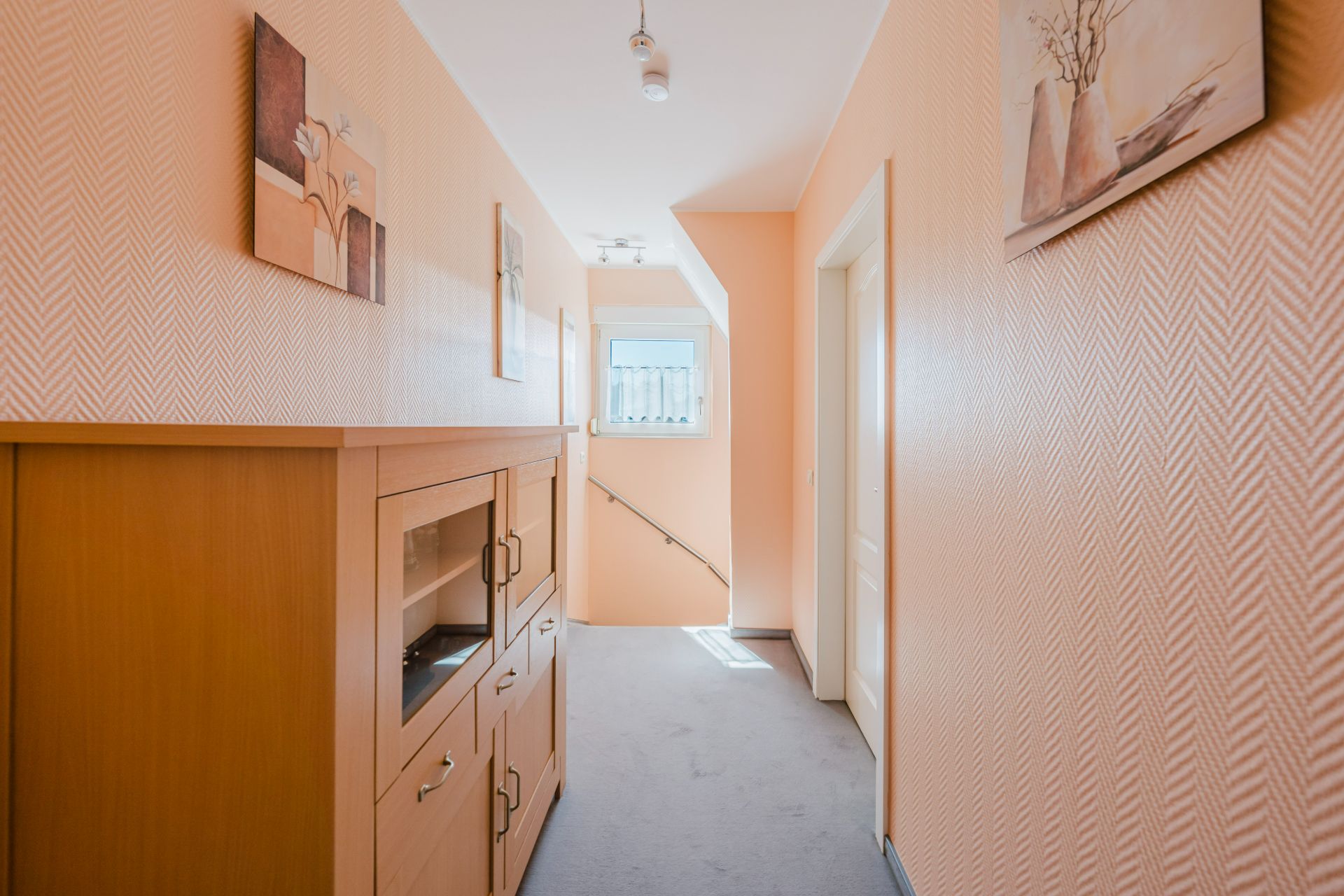 Heller Flur mit gemusterten Wänden, Holzschrank, Bildern und Treppe am Ende.