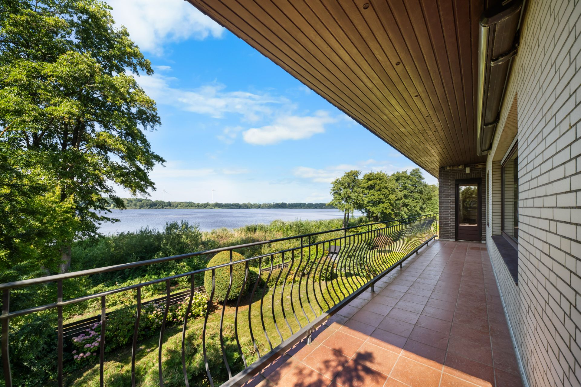 Großer Balkon mit Fliesenboden und Blick auf Garten und See, umgeben von Bäumen.