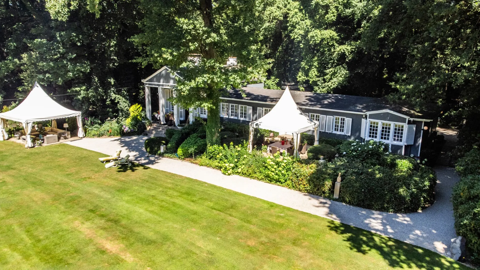 Langes Wohnhaus mit weißen Pavillons im Garten, umgeben von Bäumen und Rasenfläche.