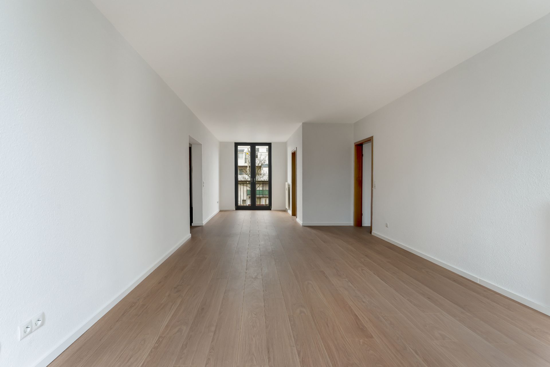 Helles, leeres Wohnzimmer mit Holzfußboden und großen Fenstern am Ende des Raumes.