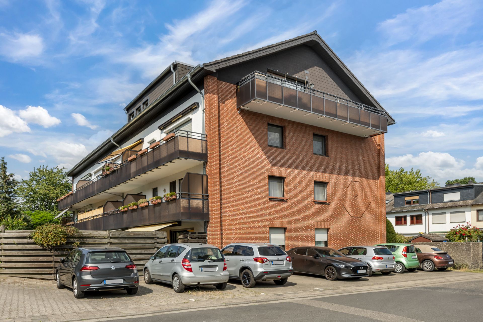 Mehrfamilienhaus mit Backsteinfassade und großem Balkon, Autos parken davor.
