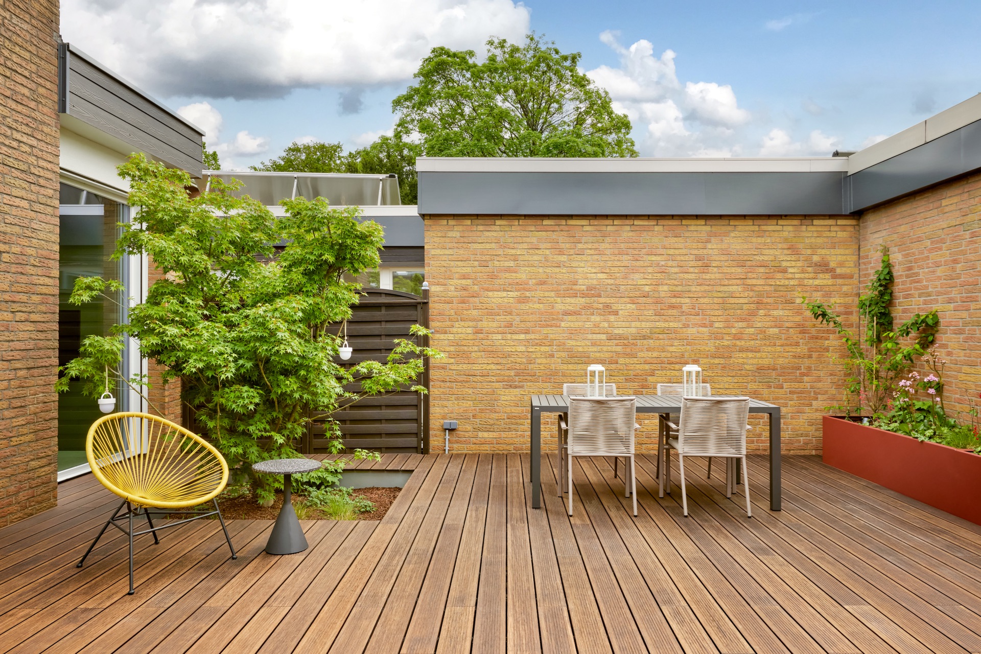 Terrasse mit Holzboden, gelbem Stuhl, Tisch, vier Stühlen und Pflanzen vor Backsteinwand.