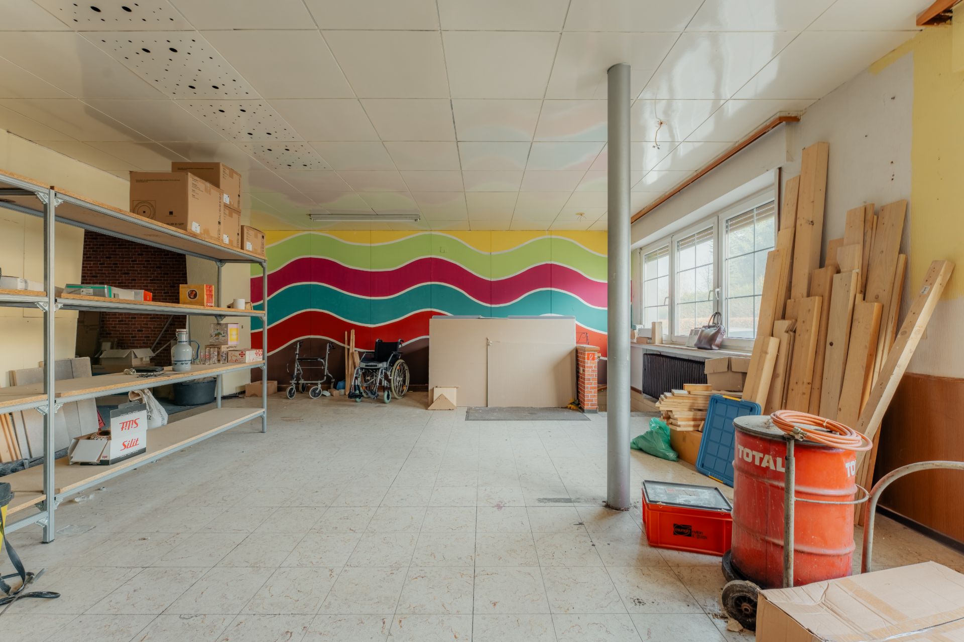 Geräumiger Abstellraum mit Regalen, Werkzeugen, Holzlatten und Rollstuhl, bunte Wandmalerei im Hintergrund.