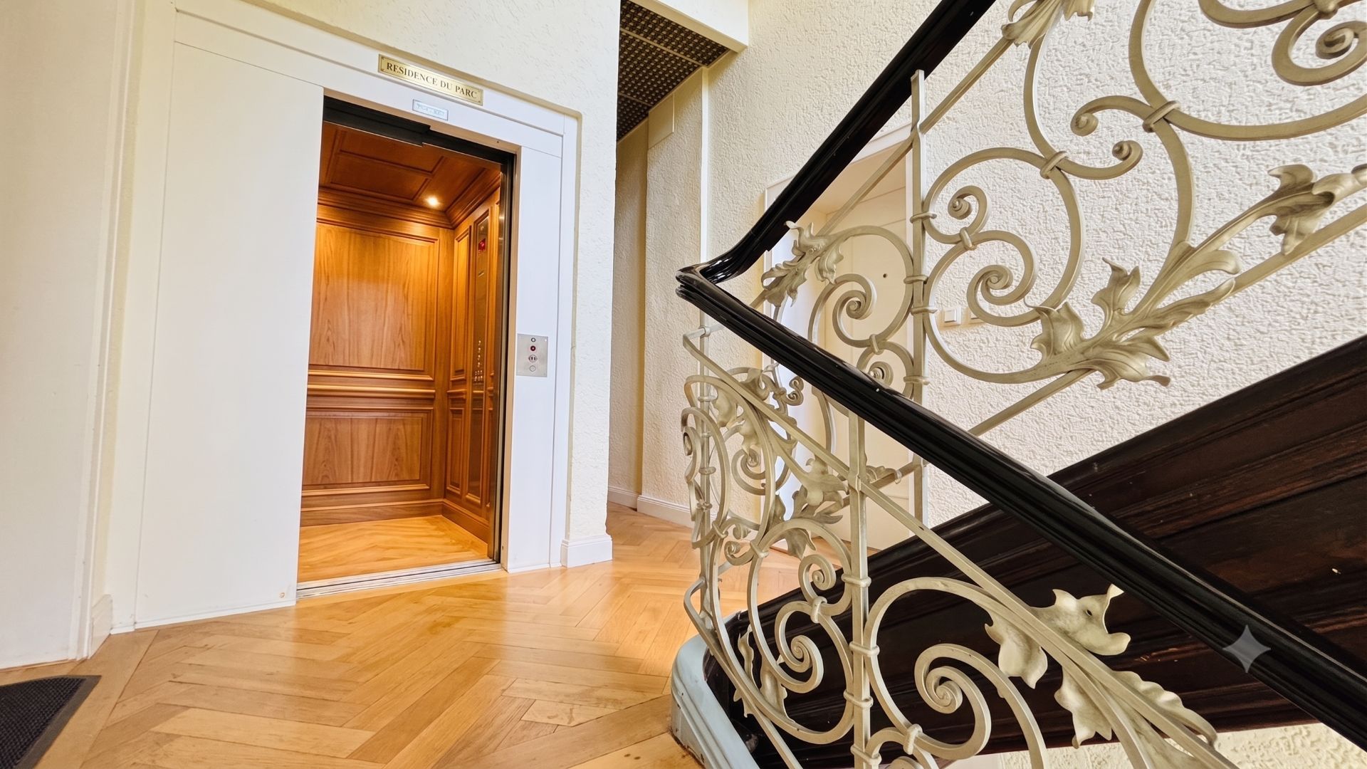 Offener Aufzug mit Holzverkleidung neben einem verzierten Treppengeländer in einem Wohnhaus.