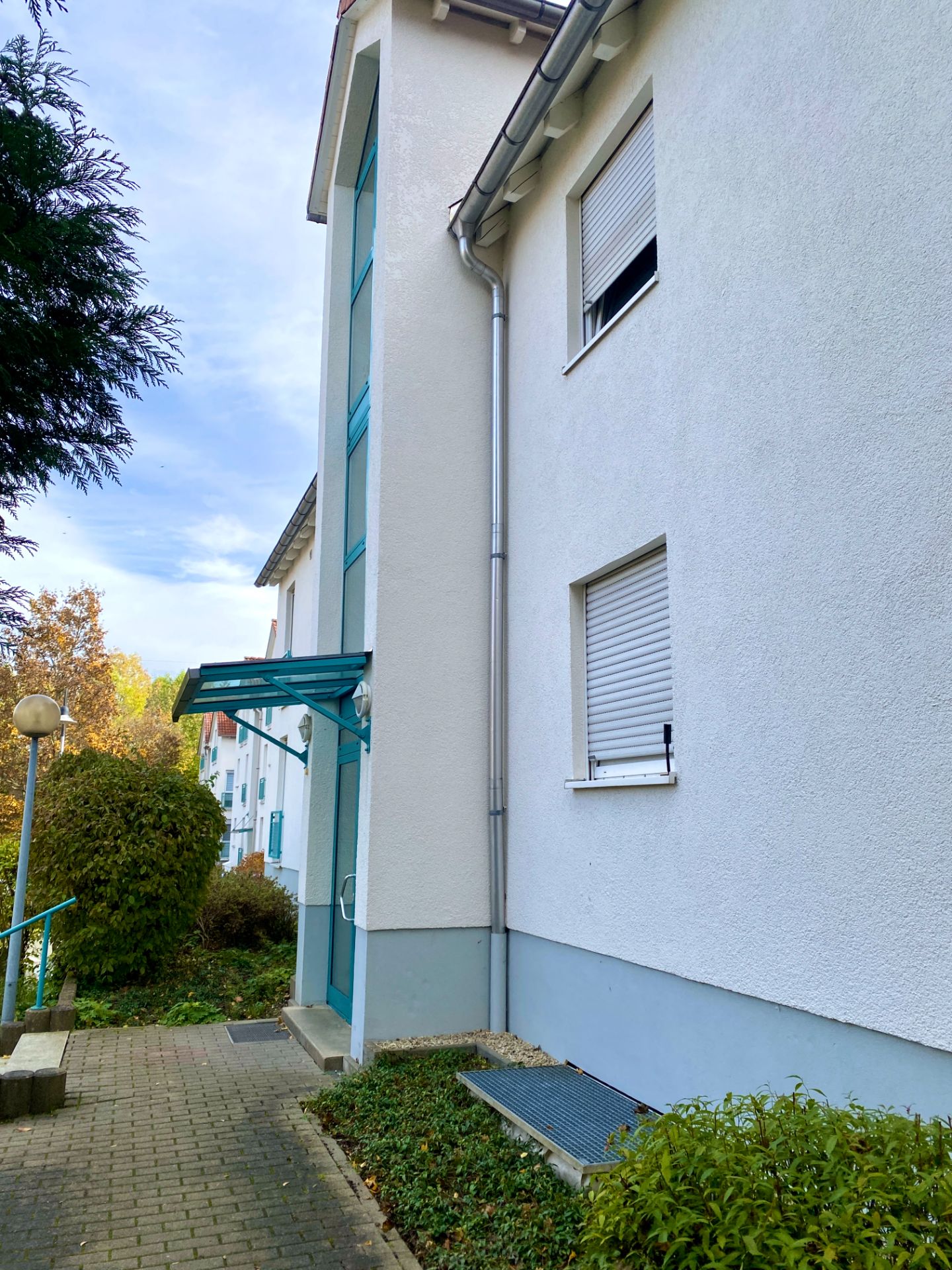 Eingang eines Wohnhauses mit weißer Fassade, Gehweg und grünen Pflanzen.