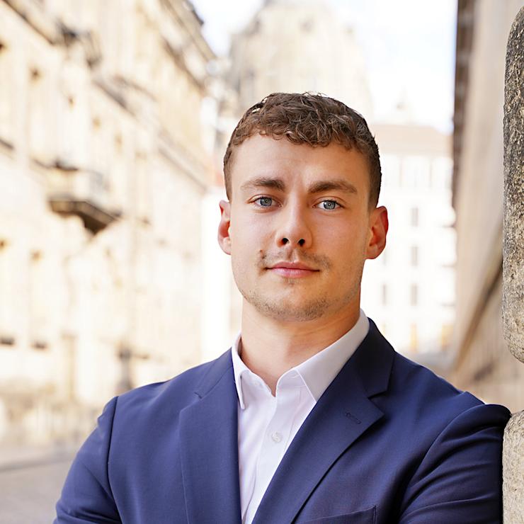Ein Mann im blauen Anzug steht mit verschränkten Armen an einer Steinwand auf einer gepflasterten Straße.