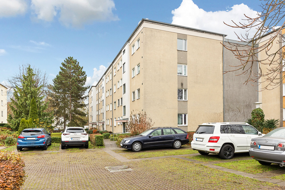 Mehrfamilienhaus mit mehreren Pkw auf einem Parkplatz und Bäumen im Hintergrund.