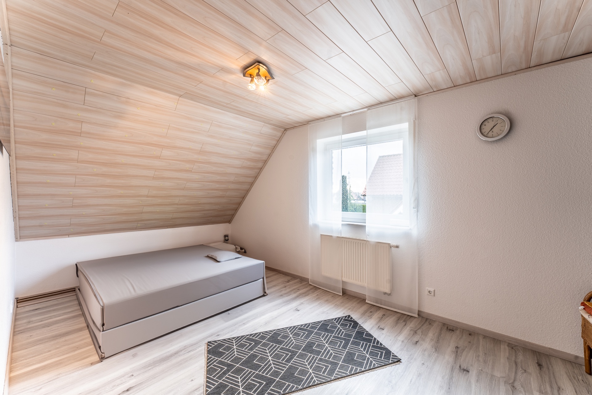 Helles Schlafzimmer mit Einzelbett, Fenster, Teppich, Wanduhr und schräger Holzdecke.