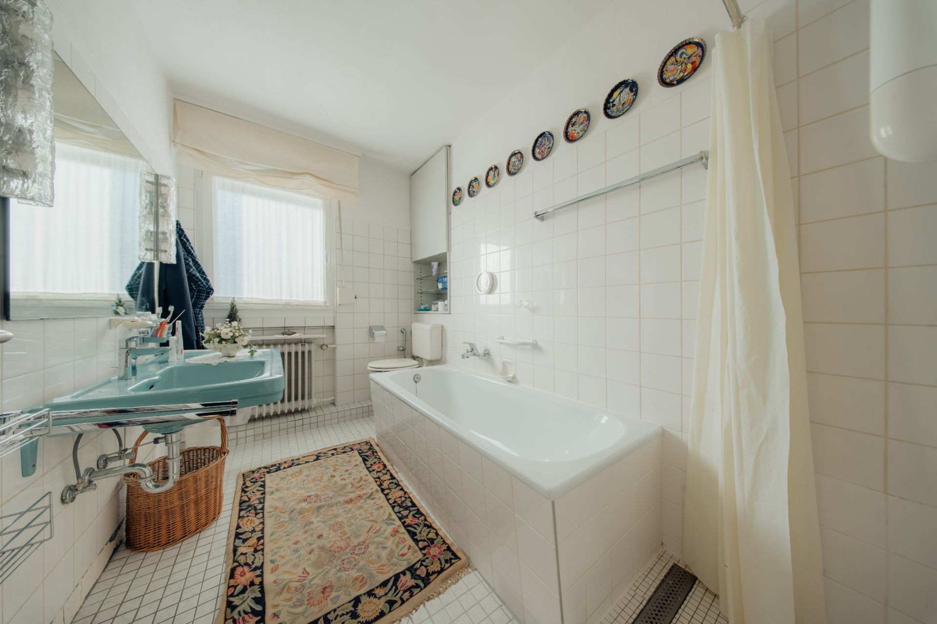 Helles Badezimmer mit Badewanne, Waschbecken, Spiegel, Fenster und Teppich auf Fliesenboden.