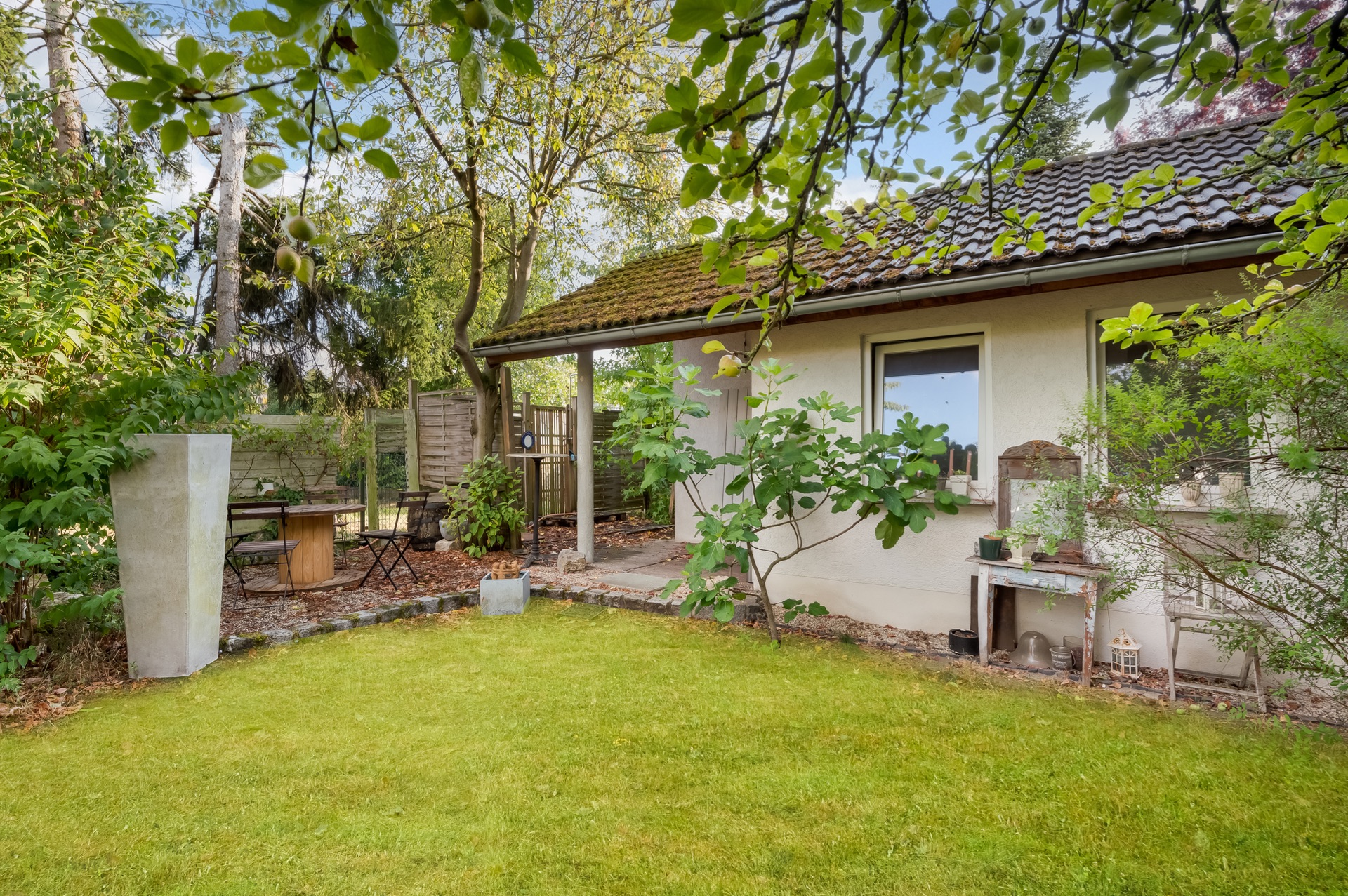 Garten mit Rasenfläche, Bäumen, Sitzbereich und Blick auf ein Haus mit zwei Fenstern.